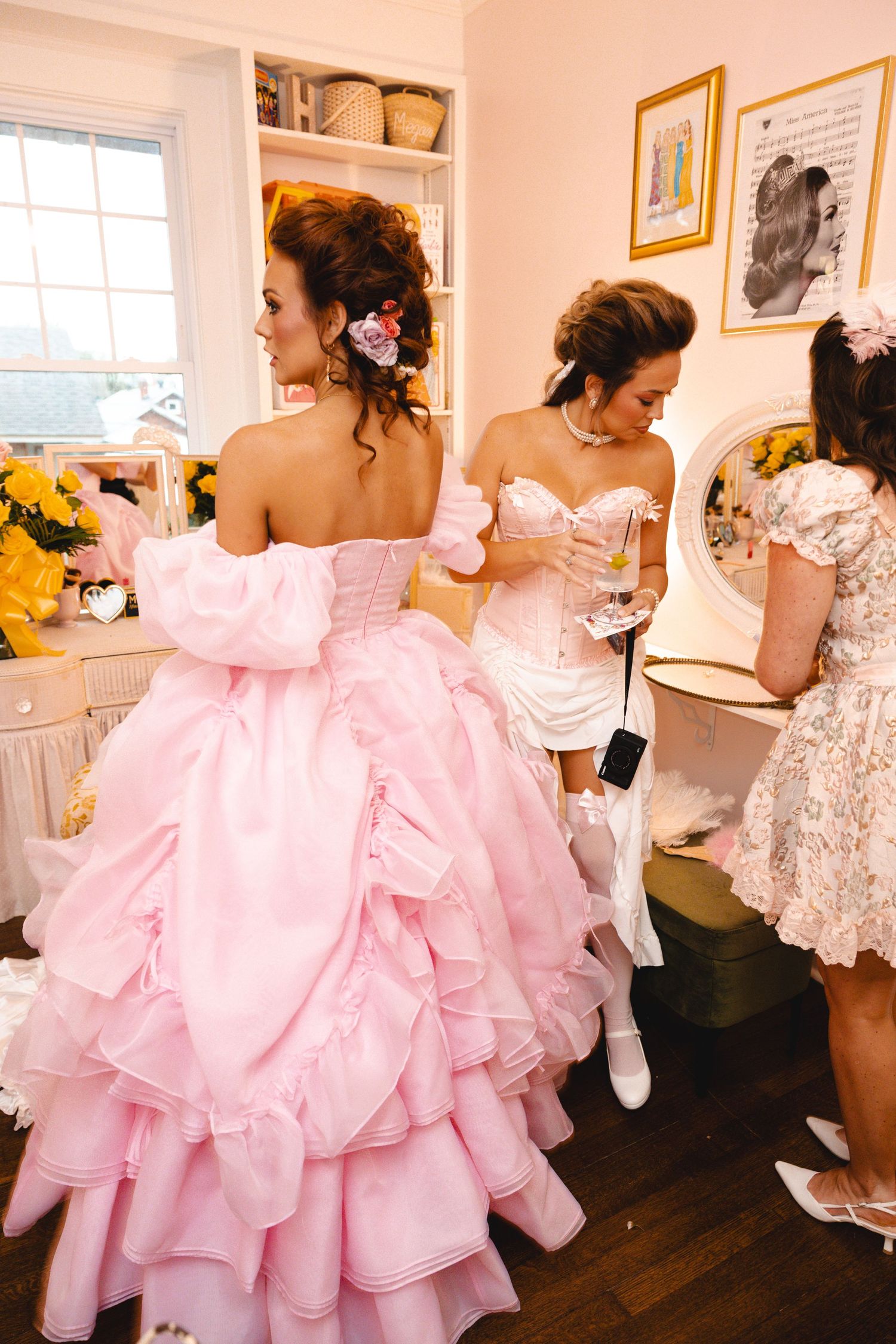Two women in elegant pink ball gowns with ruffled skirts prepare in a decorated dressing room.