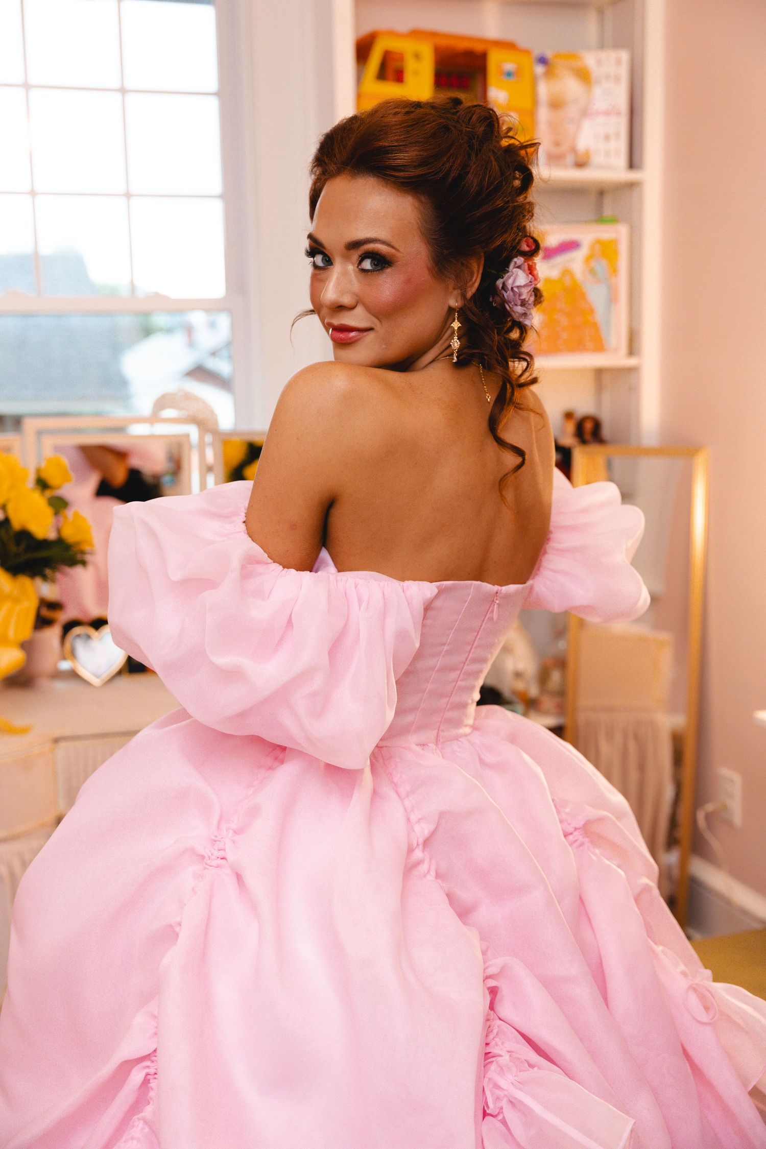 Woman in elegant pink ballgown with off-shoulder puffy sleeves poses indoors, showcasing her updo hairstyle and glamorous makeup.