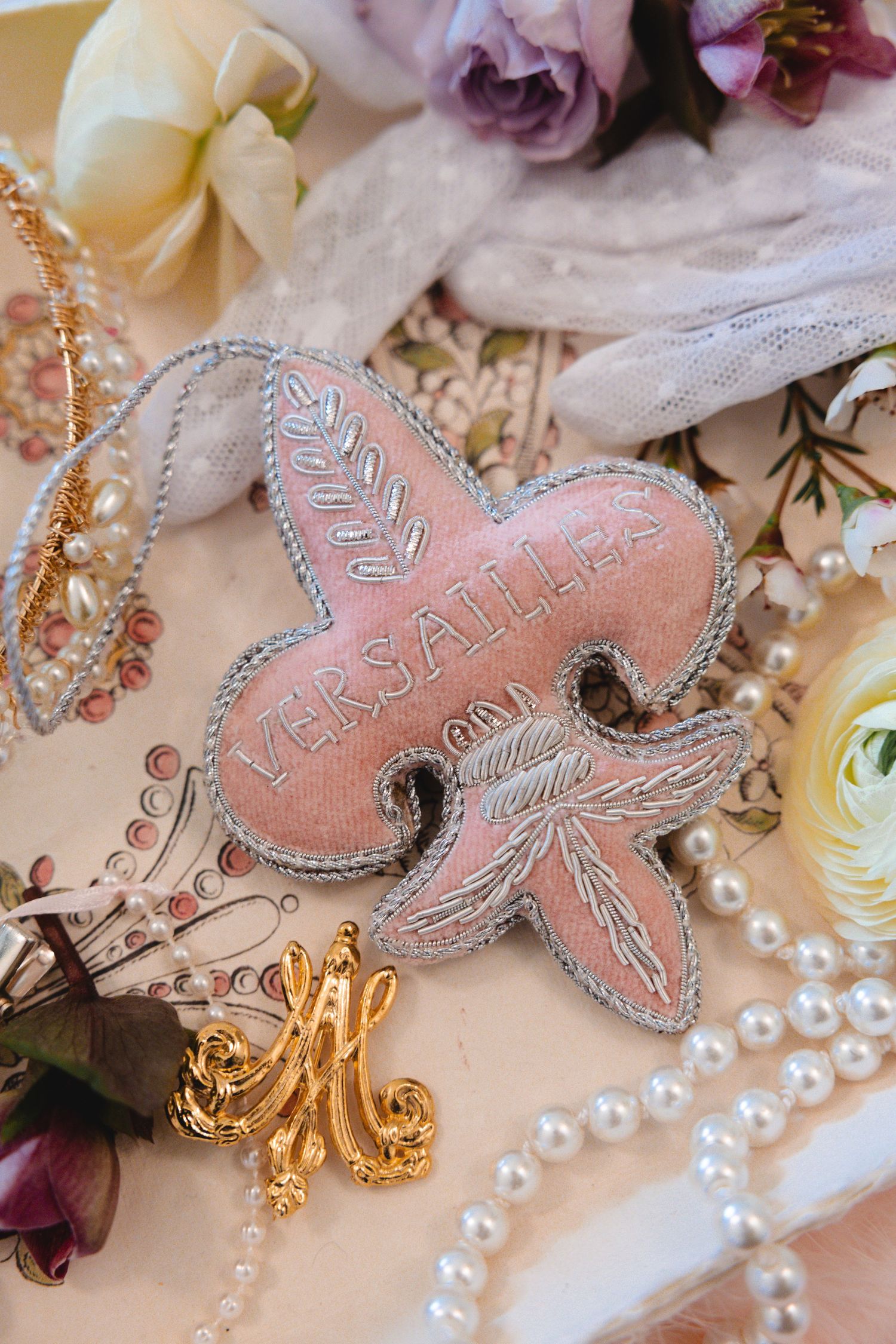 Pink fleur-de-lis embroidered Versailles patch surrounded by pearls, gold brooch, and roses on floral background.