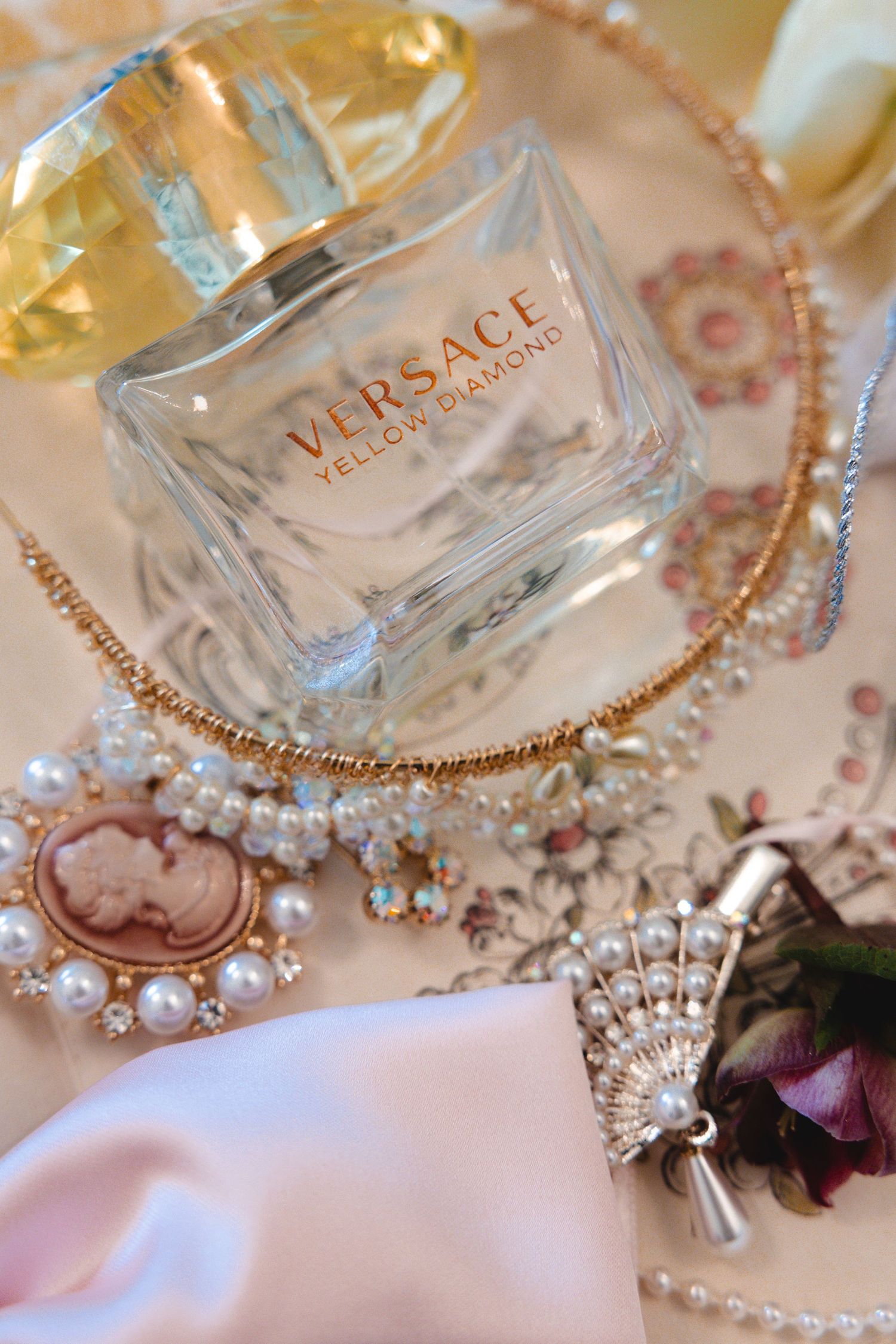 Versace Yellow Diamond perfume bottle surrounded by elegant pearl jewelry, gold necklaces, and a cameo brooch on a floral surface.
