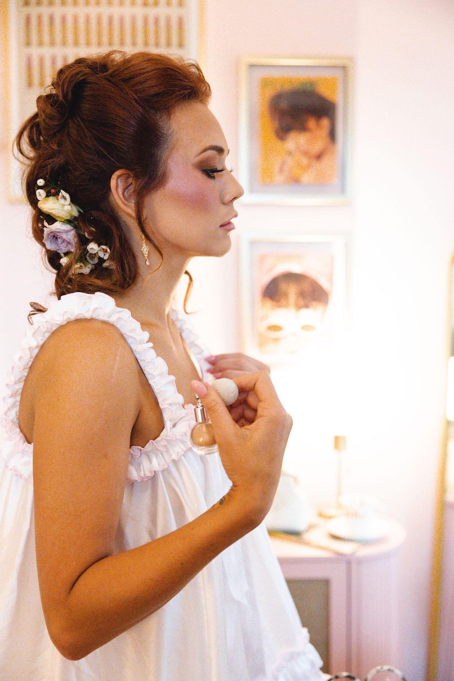 Bride in white dress applying perfume, elegant updo with flowers, soft makeup, framed artwork in background.