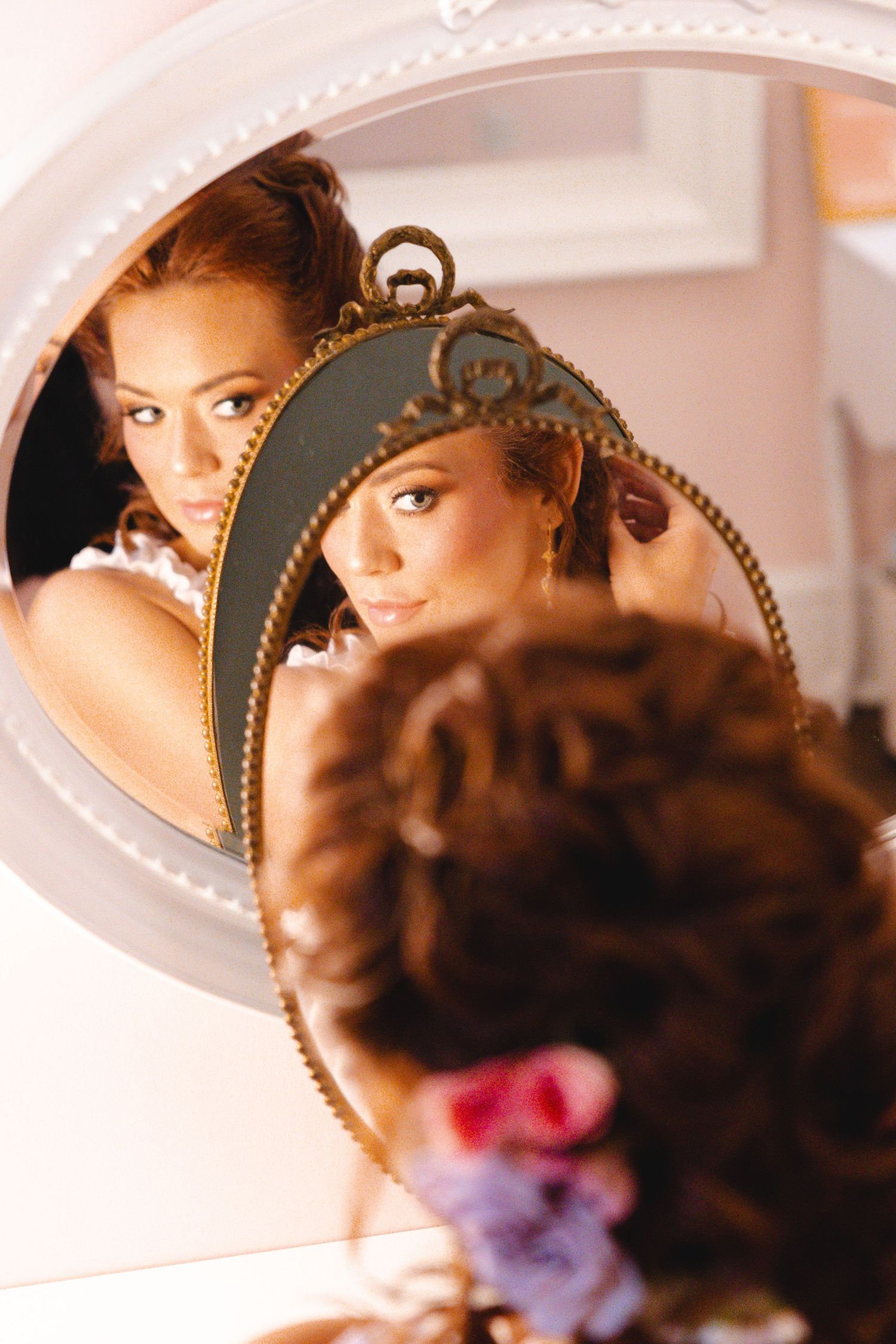 Bride with curly hair and floral accessories gazes at her reflection in an ornate gold-framed mirror.