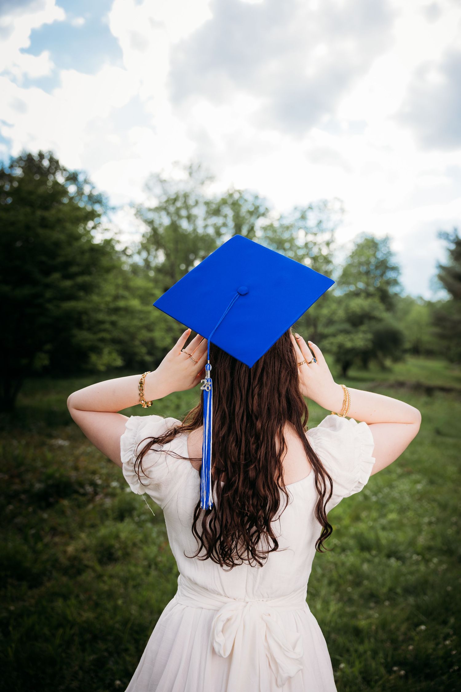 Young woman in white dress holding blue graduation cap outdoors in green field, viewed from behind.