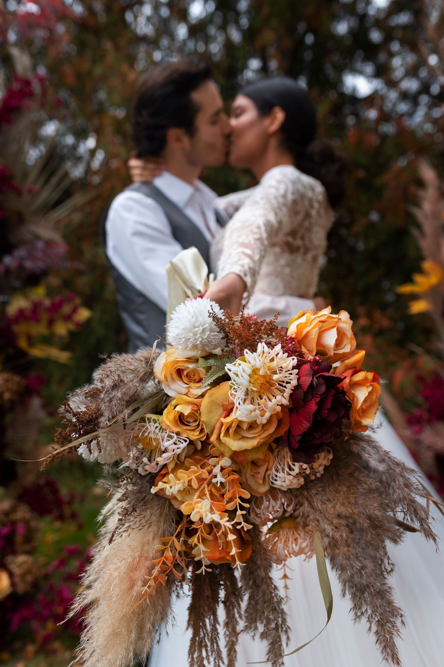 Bride and groom kissing behind a vibrant autumn wedding bouquet of orange roses and dried flowers.