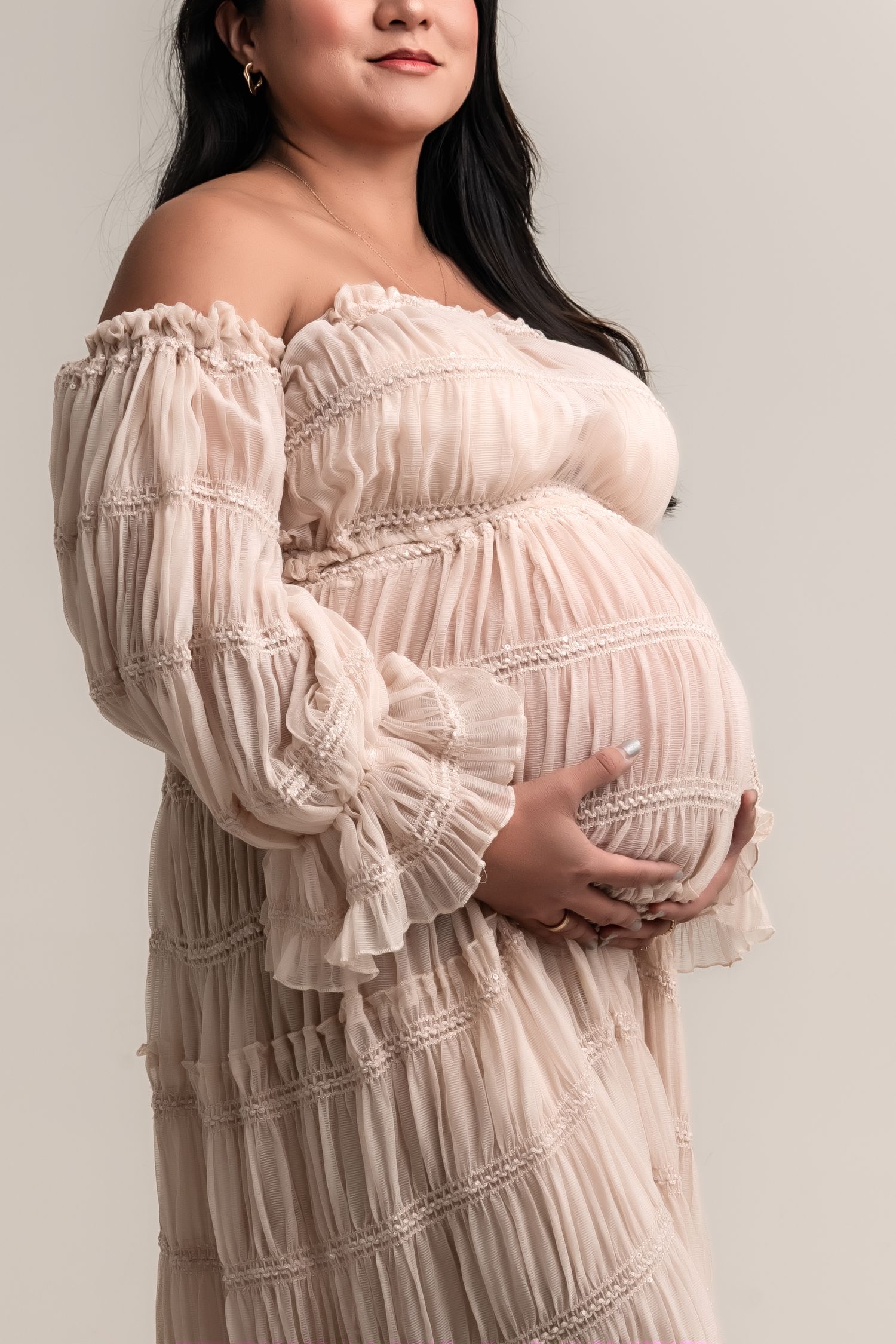 Pregnant woman in off-shoulder blush ruffle dress cradling baby bump for maternity photography studio portrait.