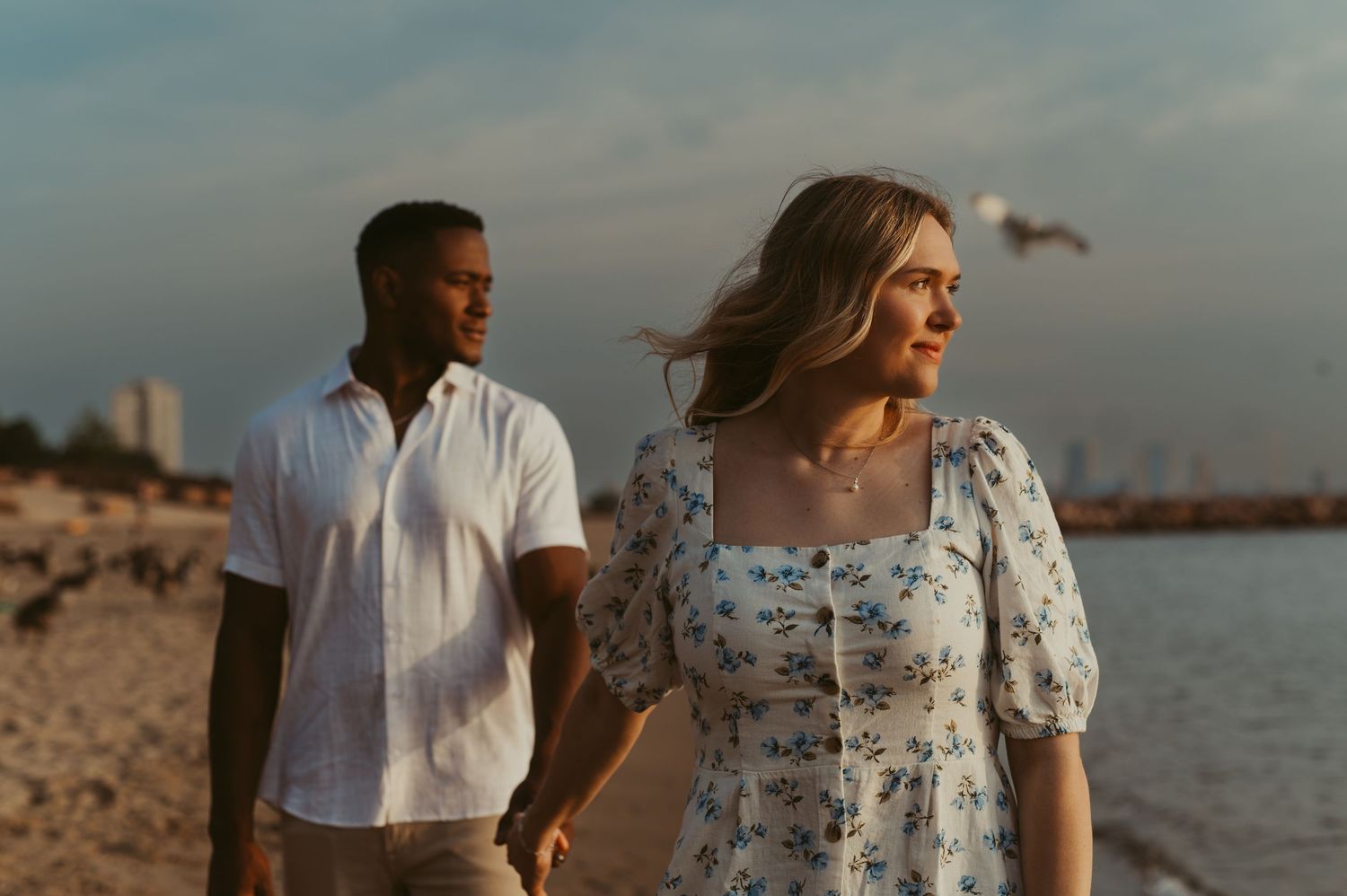 Couple walking on beach at sunrise, woman in floral dress and man in white shirt holding hands by Lake Michigan in Milwaukee.