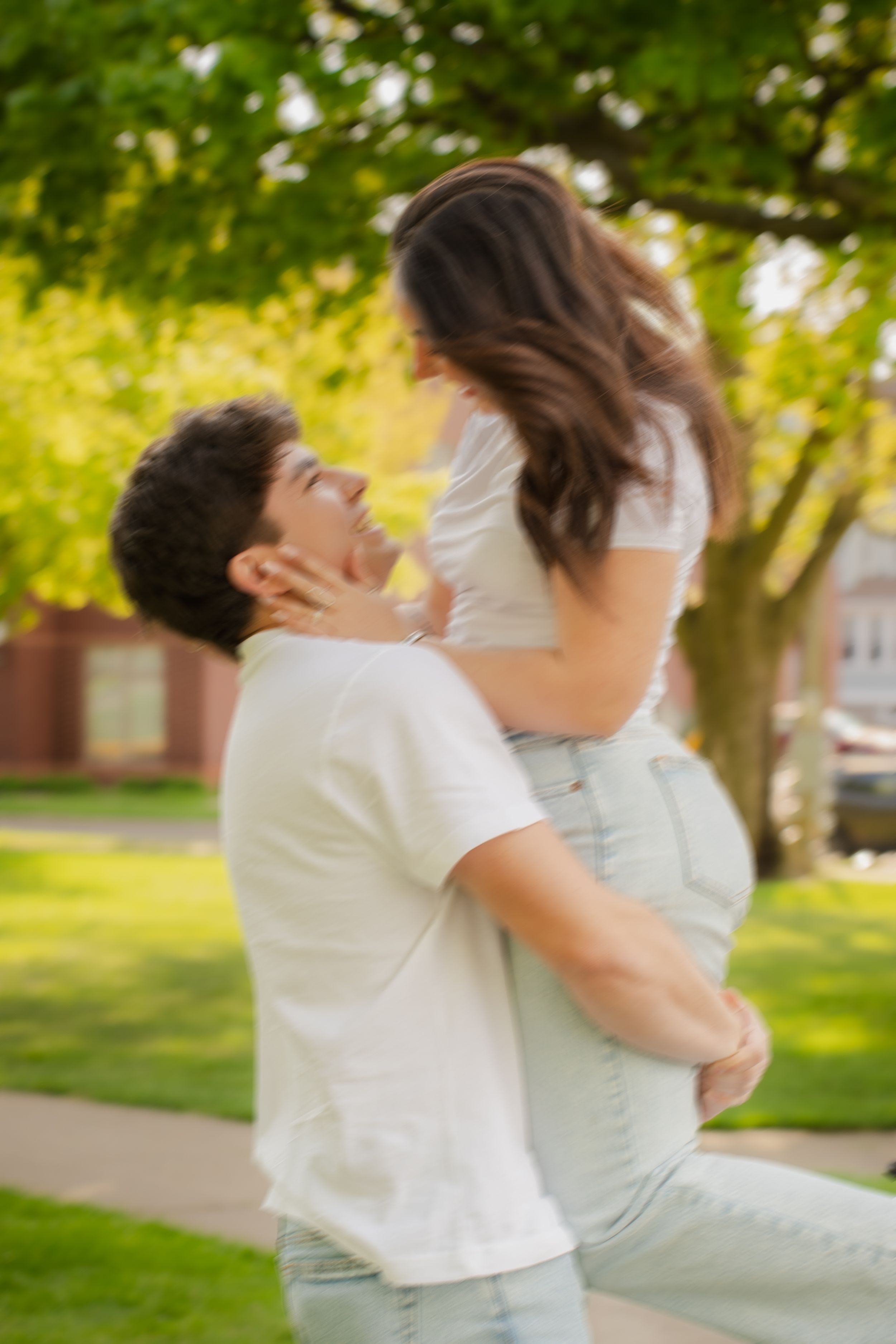 A man lifts a woman joyfully in a sunny park, both wearing white tops and jeans, sharing a romantic moment.