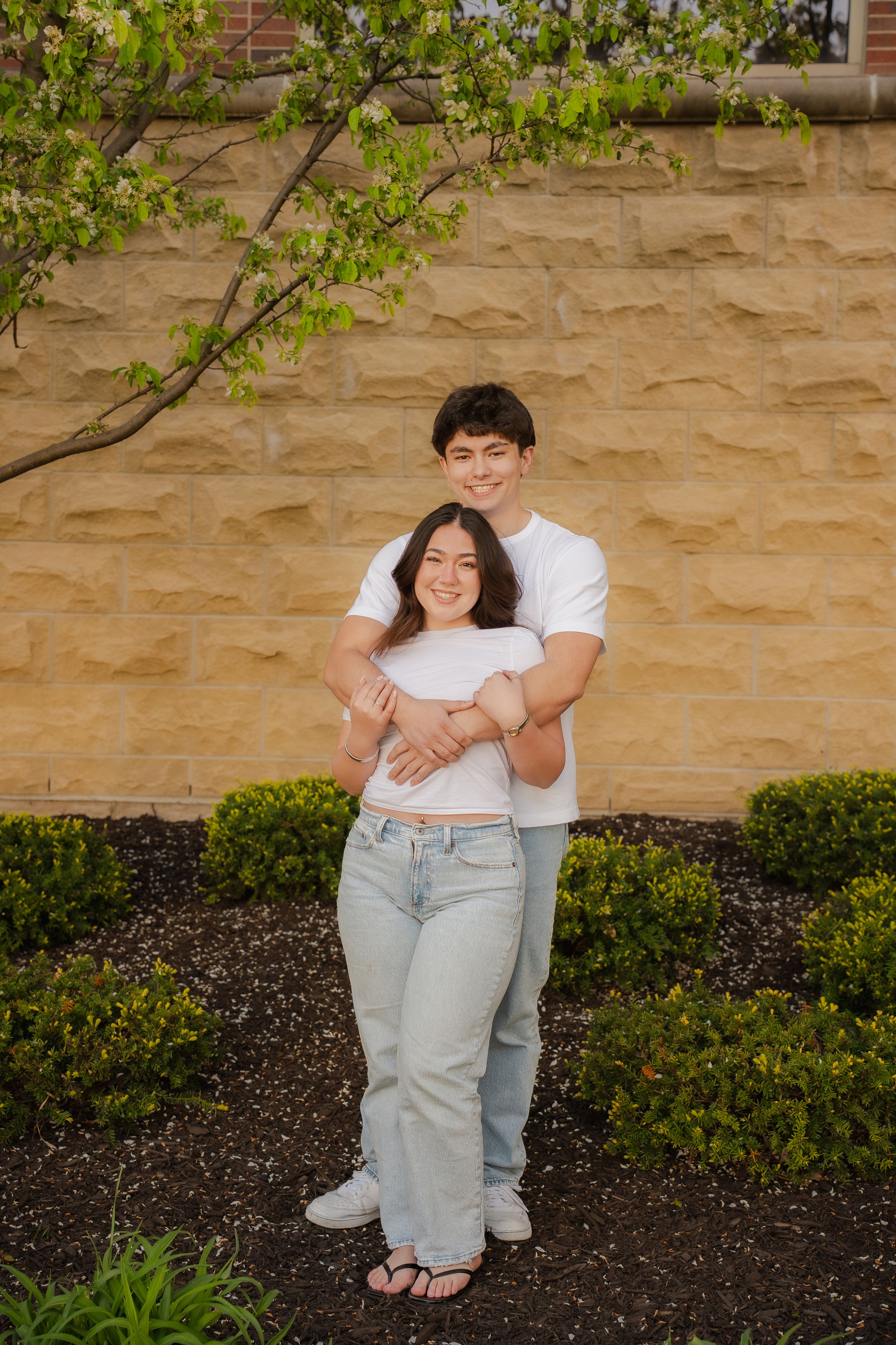 A young couple in matching white shirts and jeans embrace warmly in front of a stone wall with blooming trees.