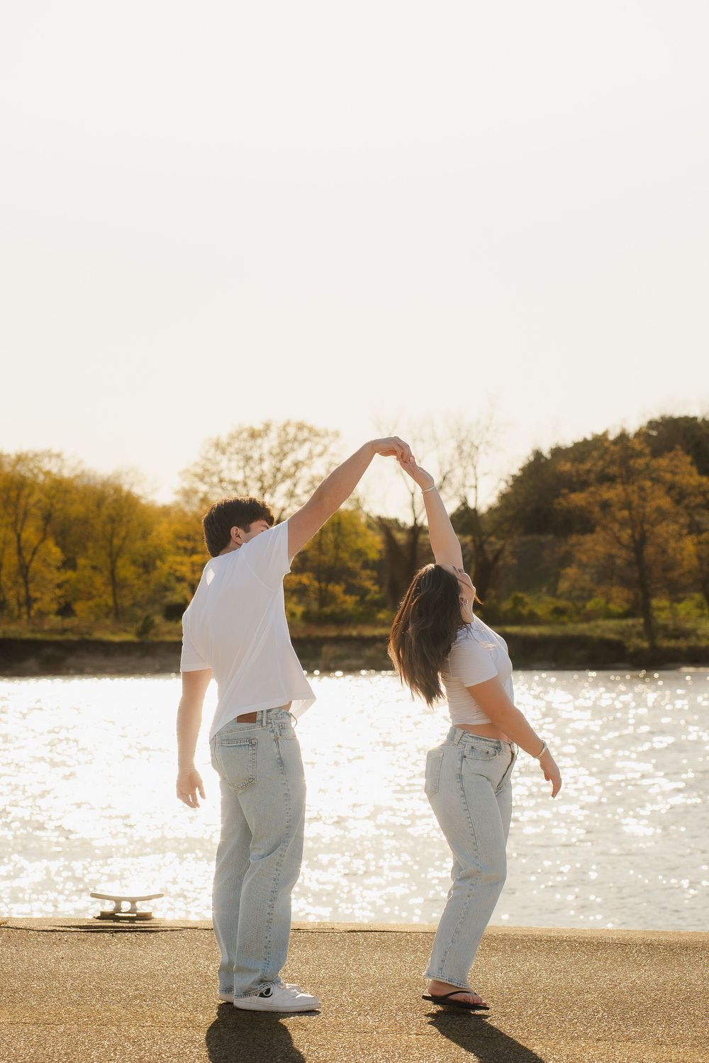 Couple dancing by a sunlit lake, holding hands raised, wearing white shirts and jeans with autumn trees in background.