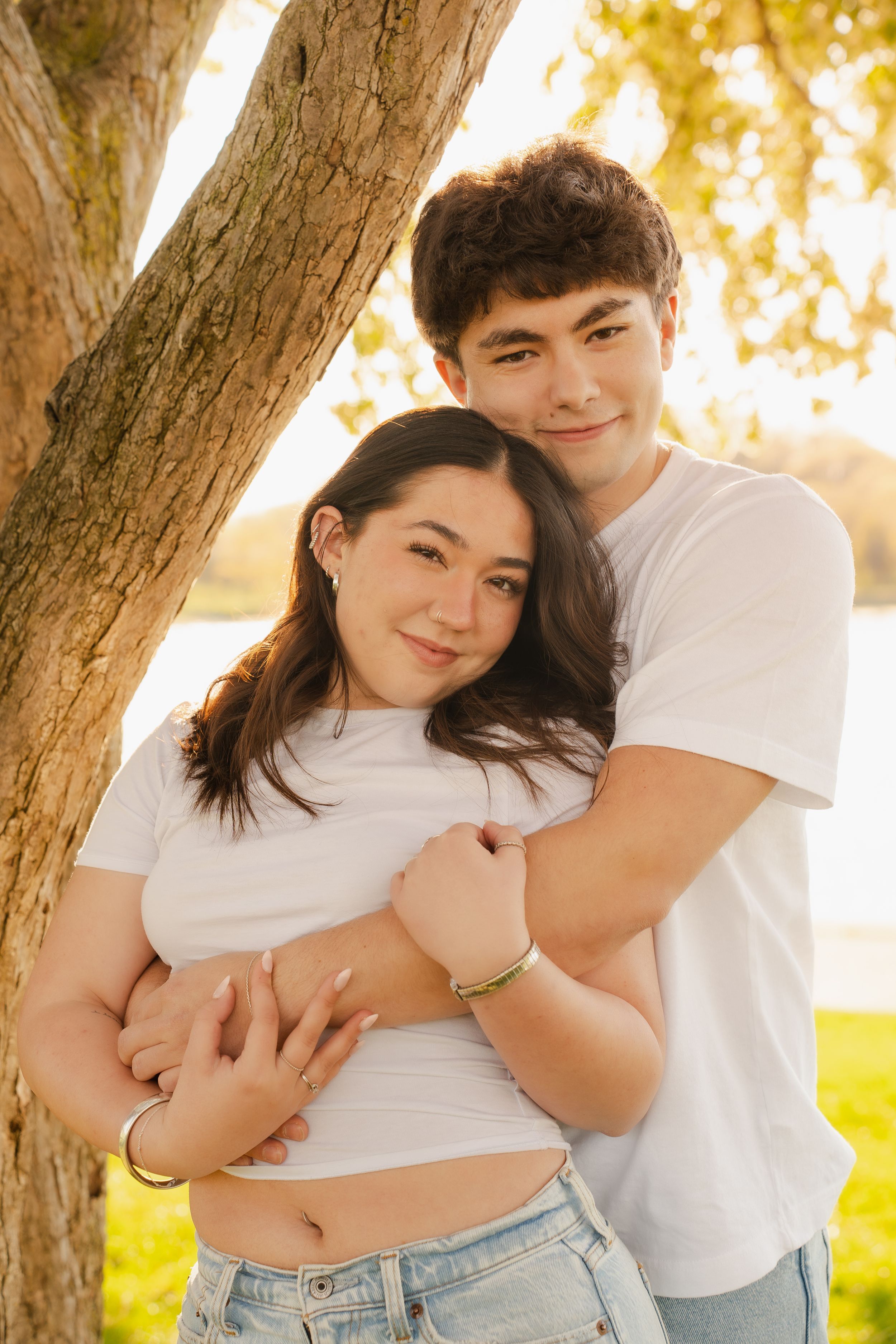 Young couple in white t-shirts embracing by a tree outdoors in warm golden sunlight.