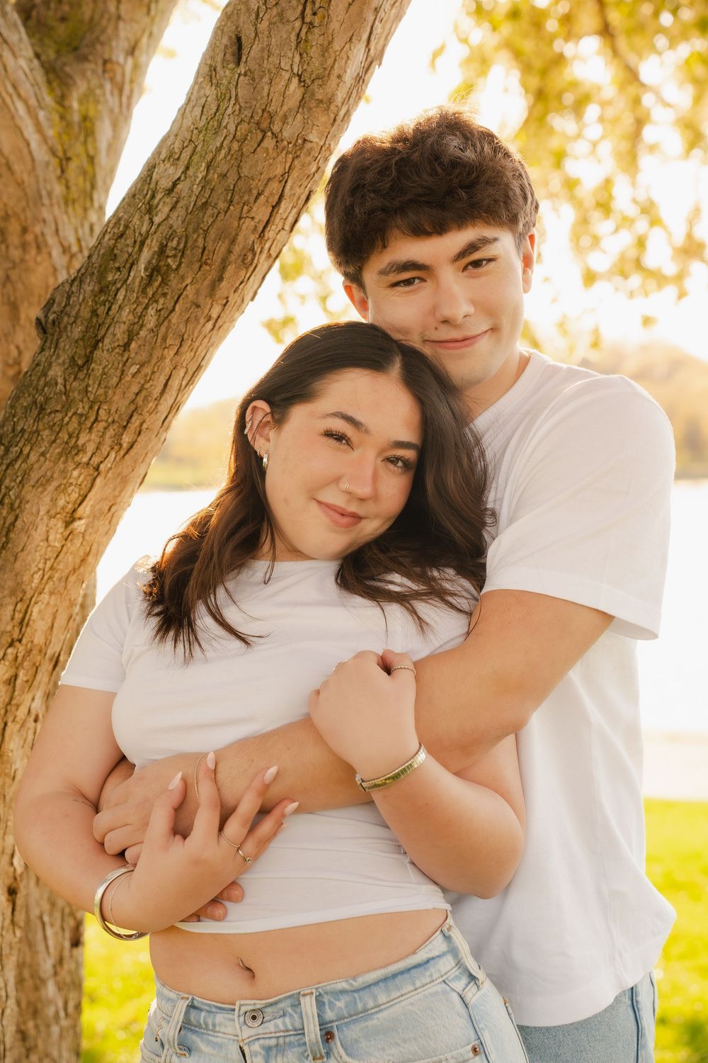 Young couple in white t-shirts embracing by a tree outdoors in warm golden sunlight.