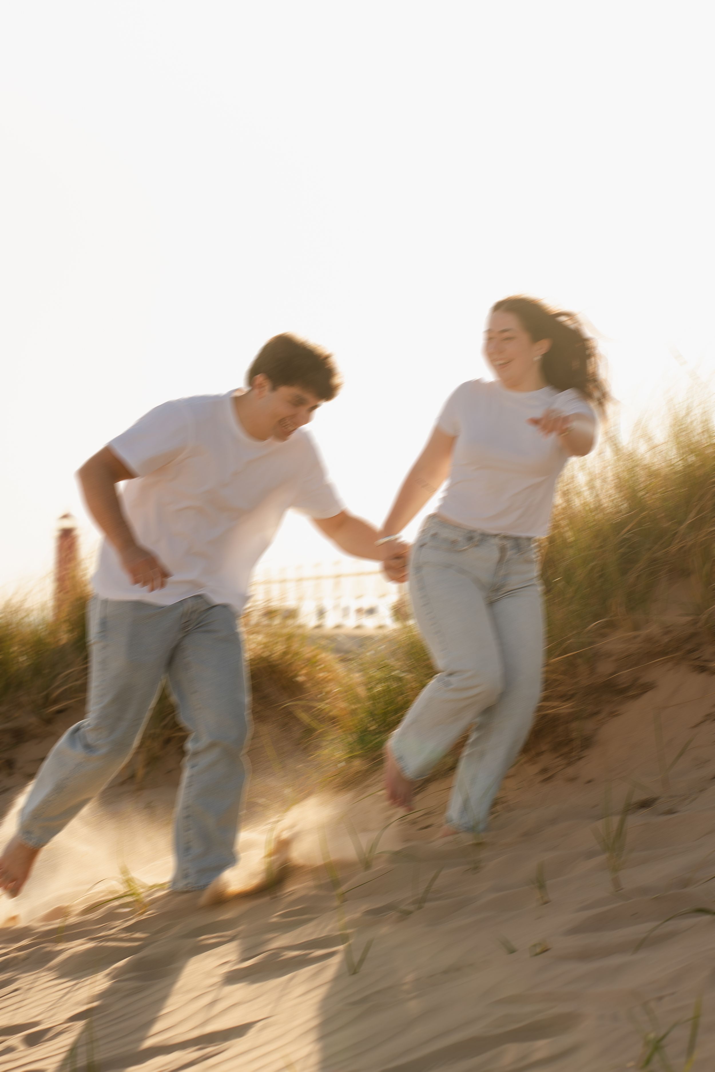 A couple in matching white tees and jeans joyfully run down a sandy beach dune at golden hour.