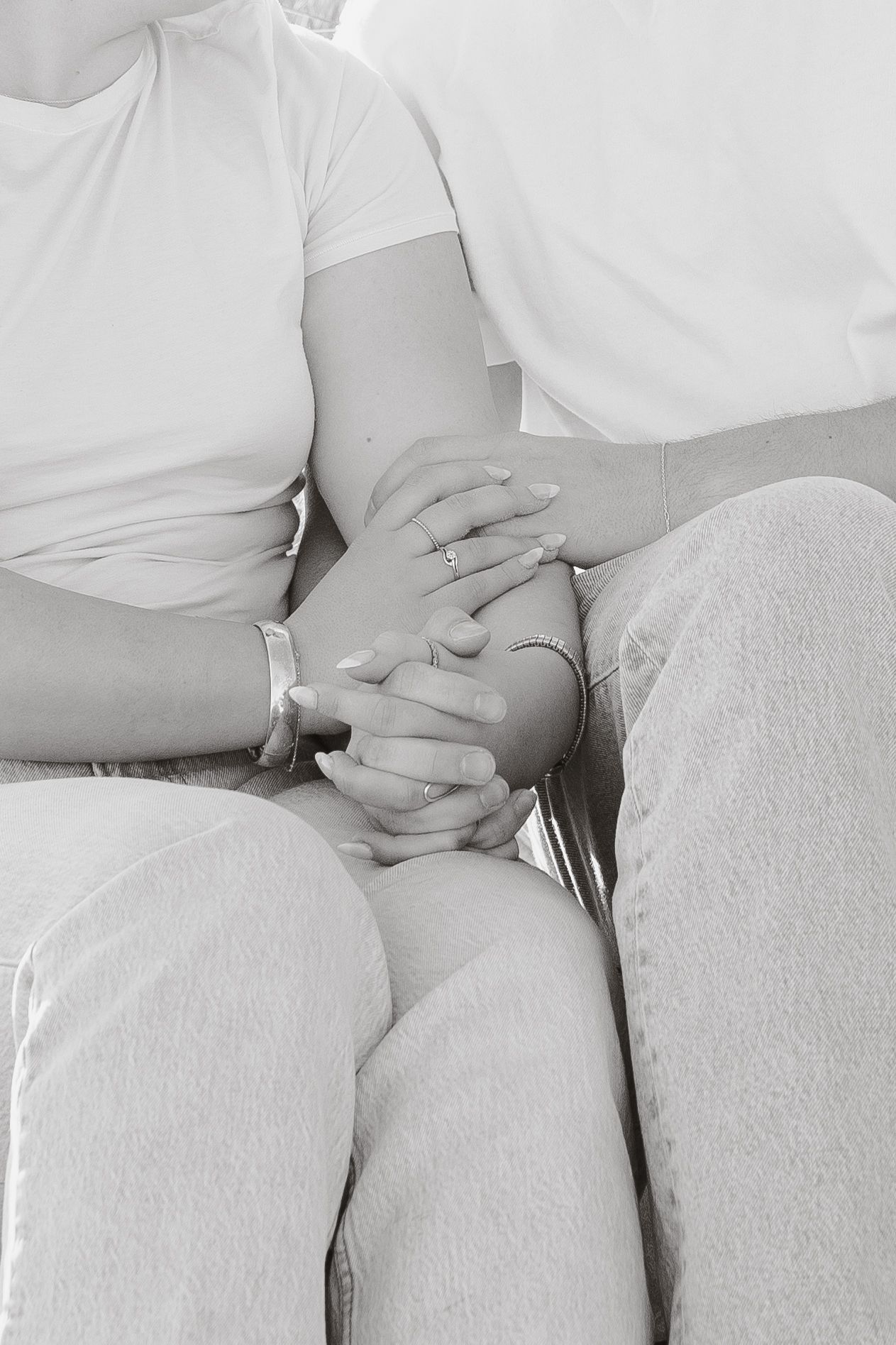 Black and white photo of a couple holding hands, showing rings and bracelets, seated together in casual white tops and jeans.
