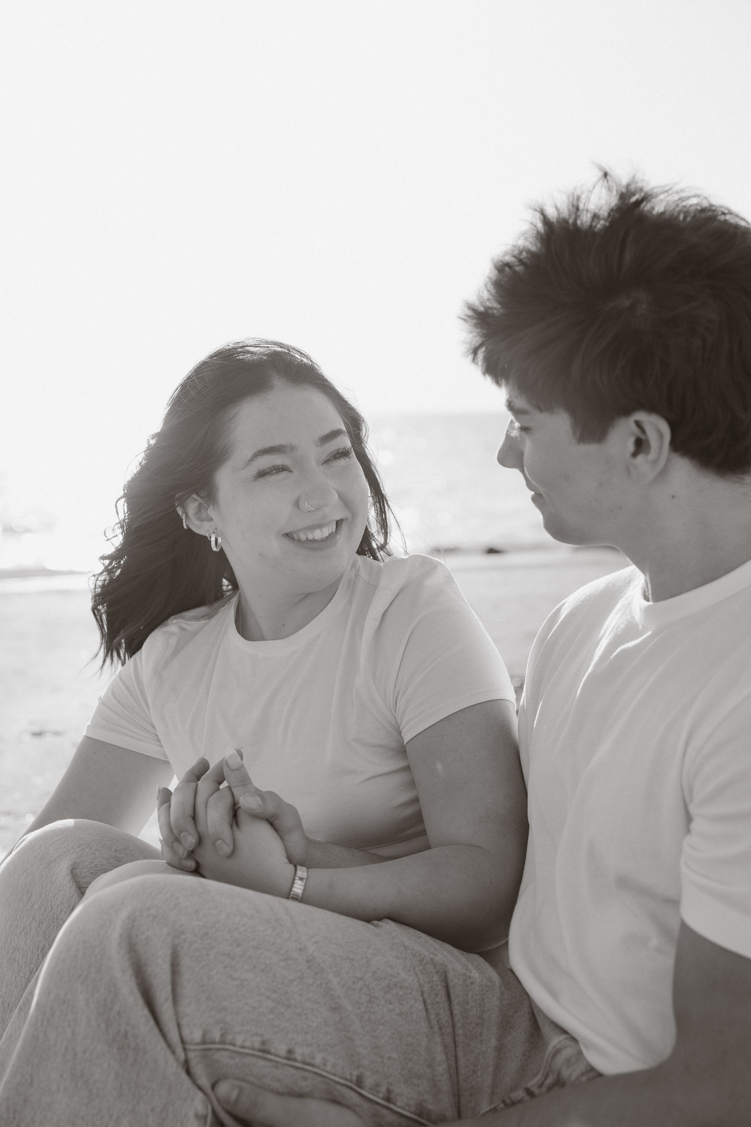 Black and white photo of a smiling couple holding hands and gazing at each other on a sunny beach.