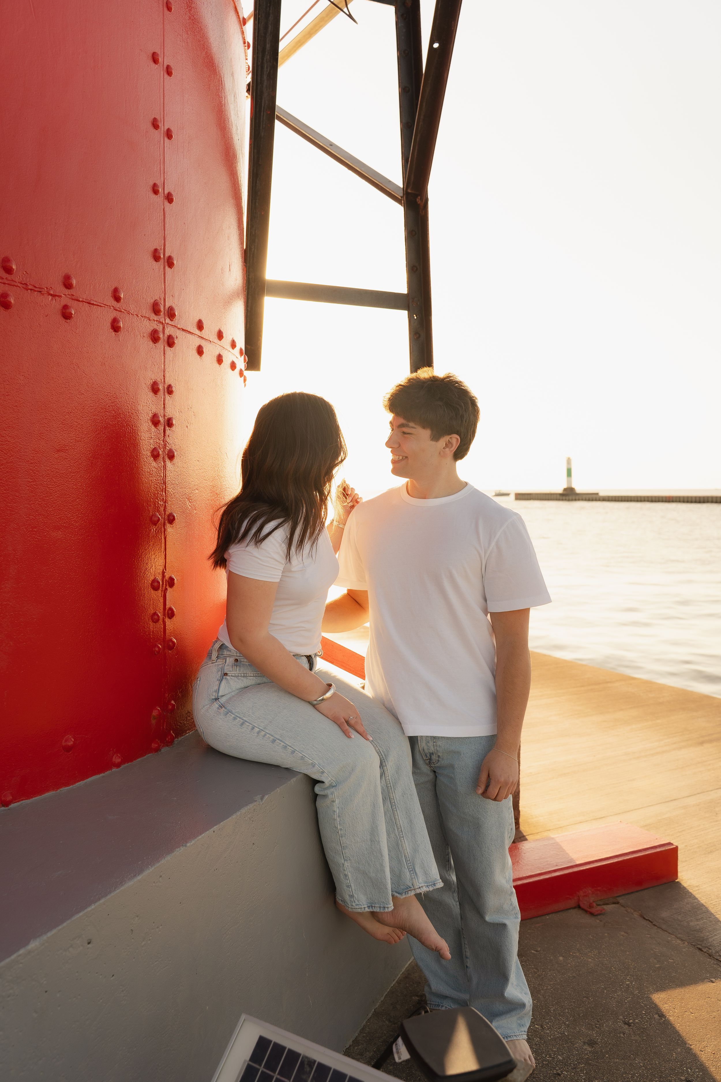 Young couple in white tees and jeans share a moment by a red lighthouse pier at golden hour sunset.