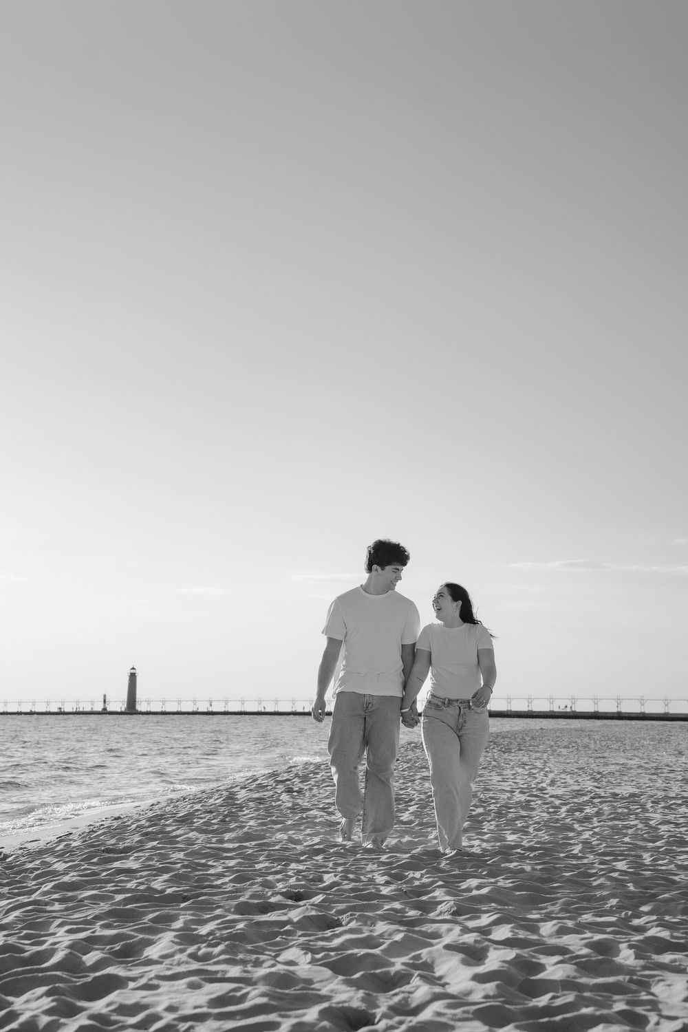 Black and white photo of a couple holding hands walking on a sandy beach with a lighthouse in the background.