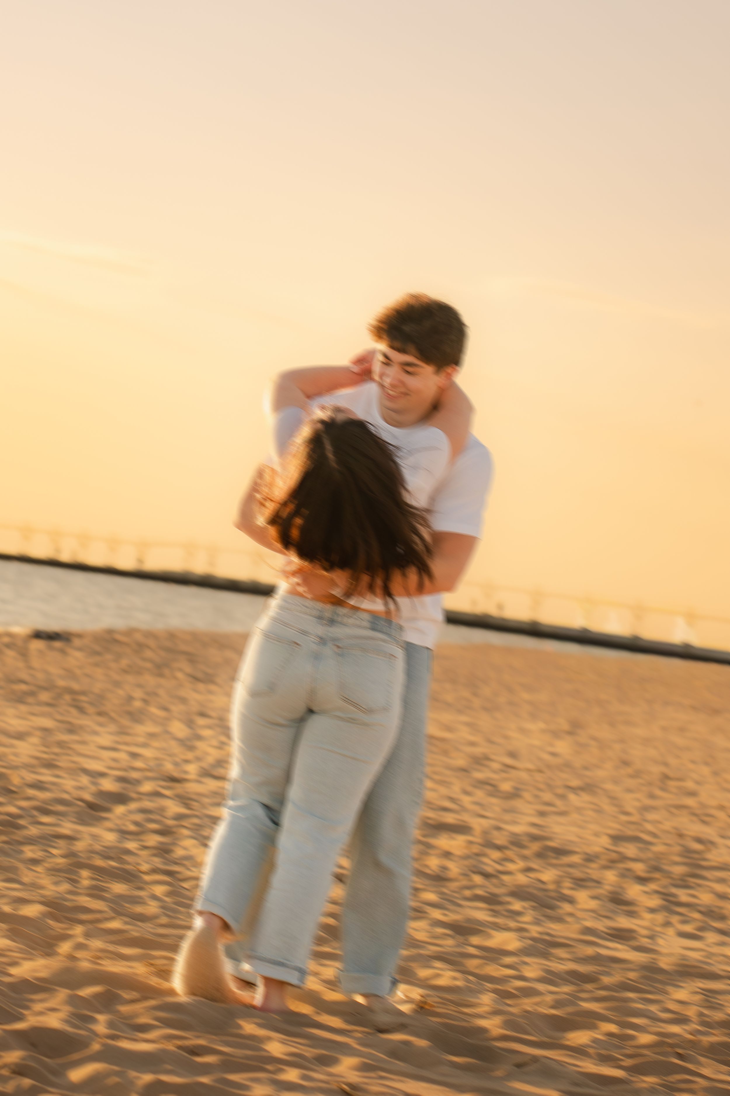 Couple embracing and spinning on a sandy beach during golden sunset, wearing white shirts and light blue jeans.