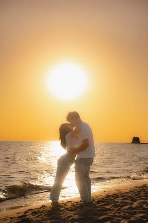 Romantic couple kissing on beach shore at vibrant orange sunset with sun glowing above the ocean horizon.