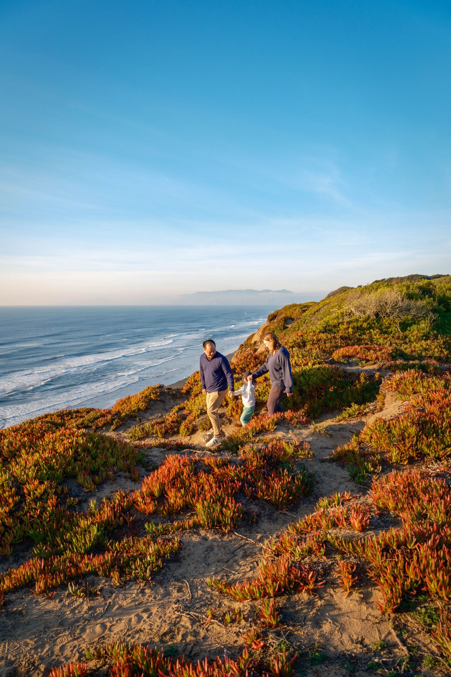 Family of three walking along a scenic coastal bluff with vibrant red ice plants and ocean views on a sunny day.