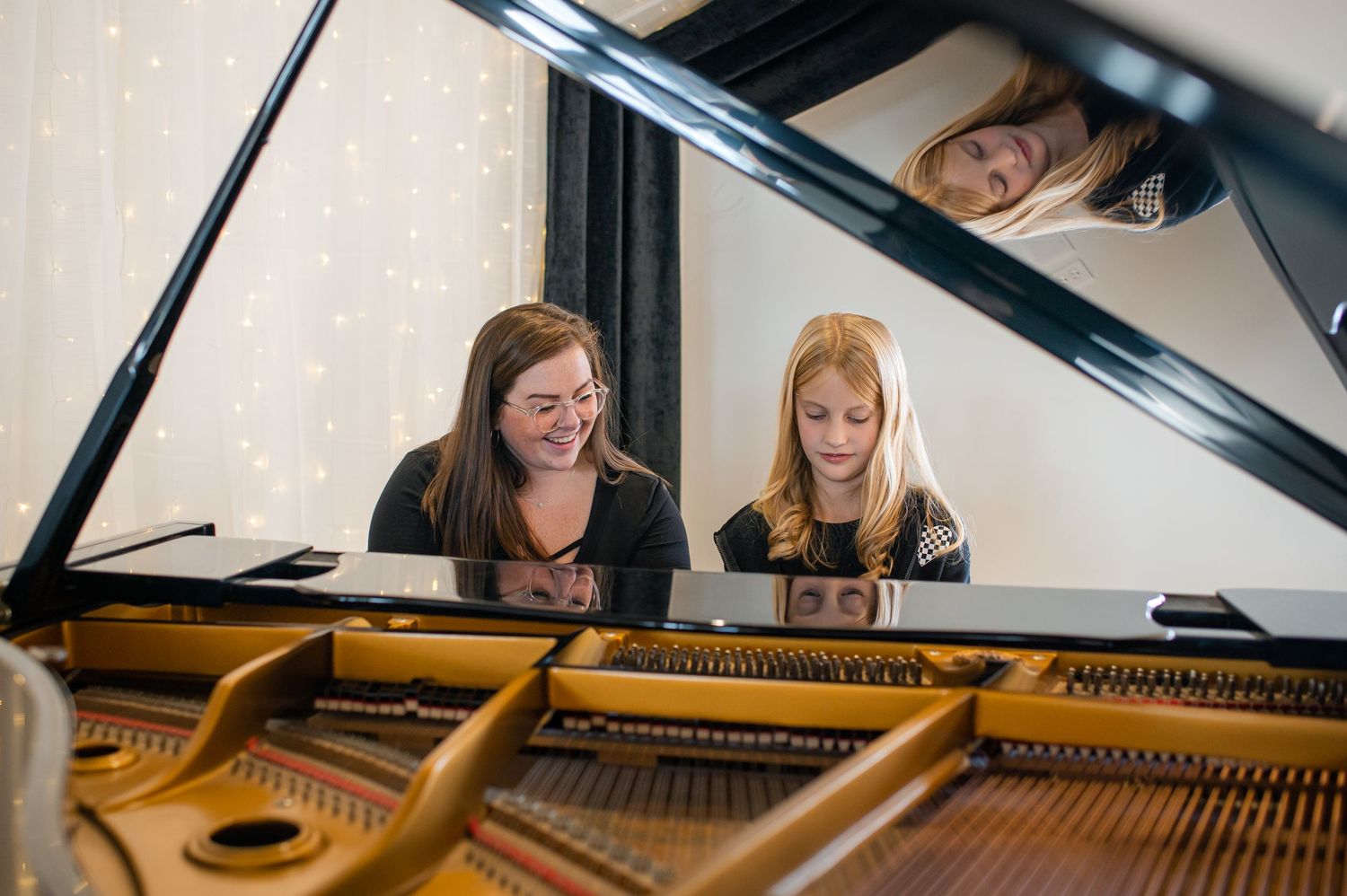 A piano teacher and young student sit together at a grand piano during a lesson, with fairy lights in the background.