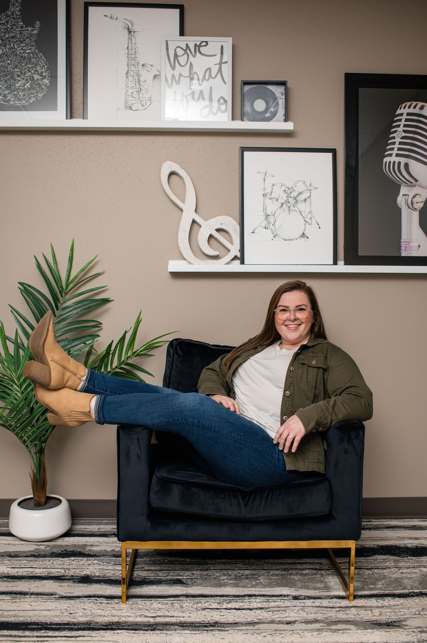 Woman smiling in navy velvet chair with music-themed wall decor, wearing jeans, green jacket, and tan boots.