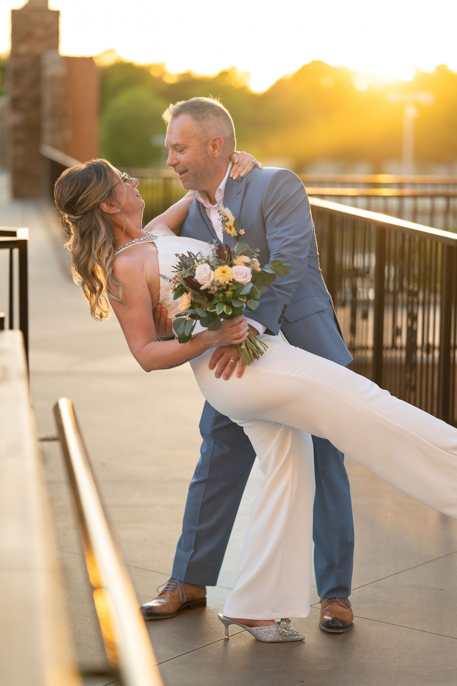 Groom dips bride in white gown at golden hour on outdoor venue bridge, she holds colorful bouquet.