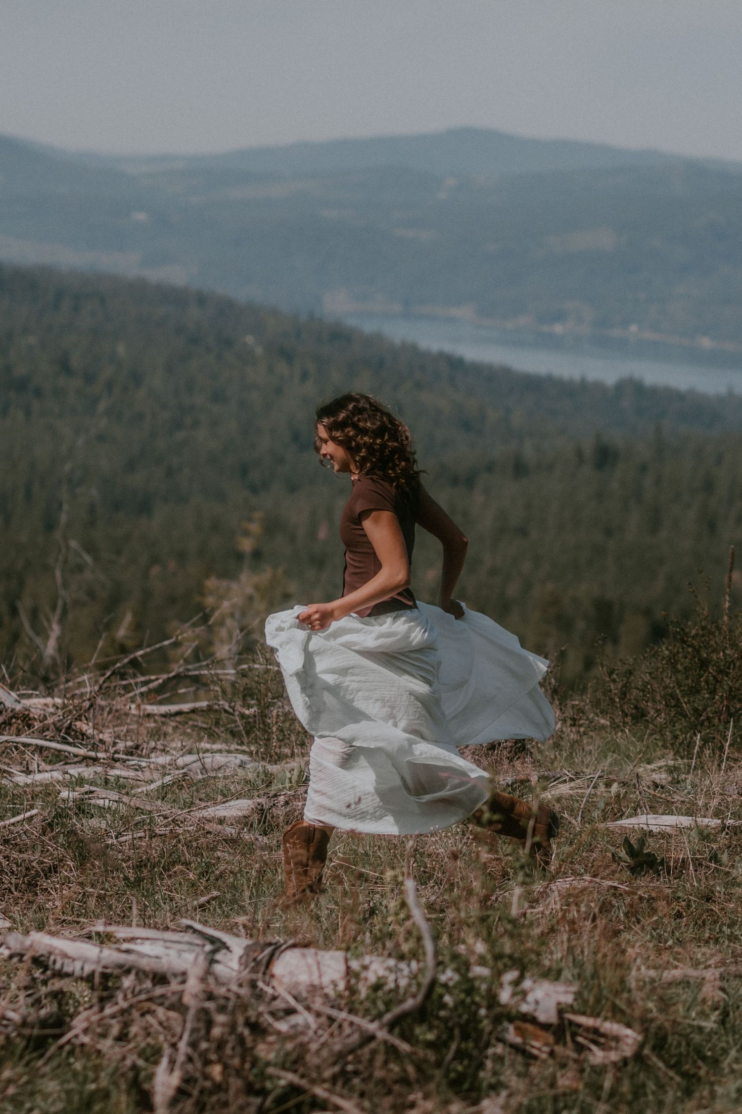 Woman in white skirt and brown top dancing on a hillside with forest and lake views in the background.