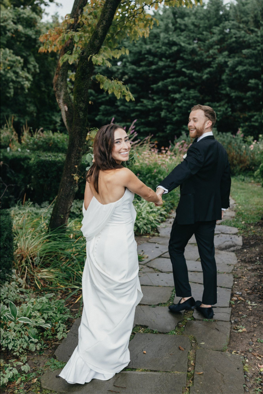 A bride and groom share a kiss outdoors near a rustic stone building, surrounded by lush greenery and overhanging tree branches.
