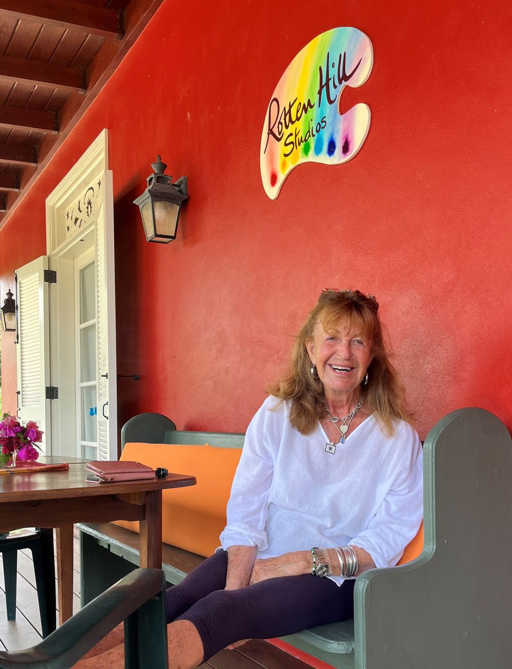 Woman smiling on colorful porch bench at Roatan Hill Studios, red wall backdrop.
