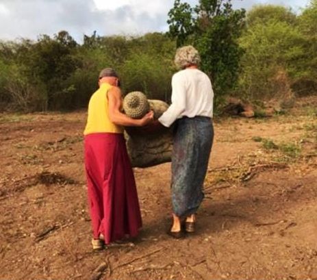 Two women in colorful skirts placing a decorative ball on a rock in a dry, earthy outdoor landscape.
