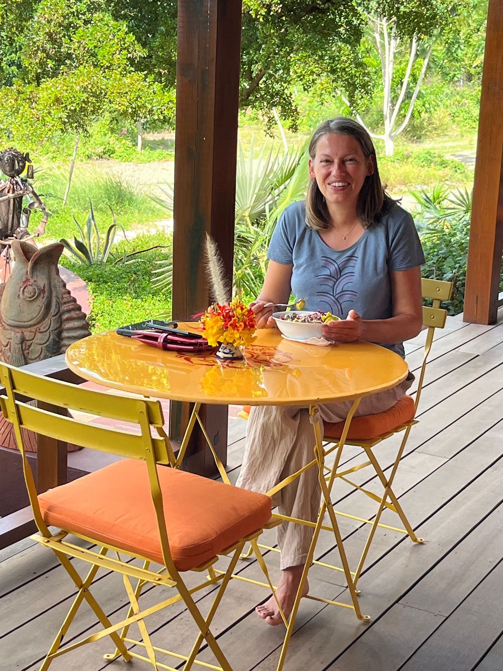 Woman smiling at yellow bistro table on wooden deck surrounded by lush green garden.