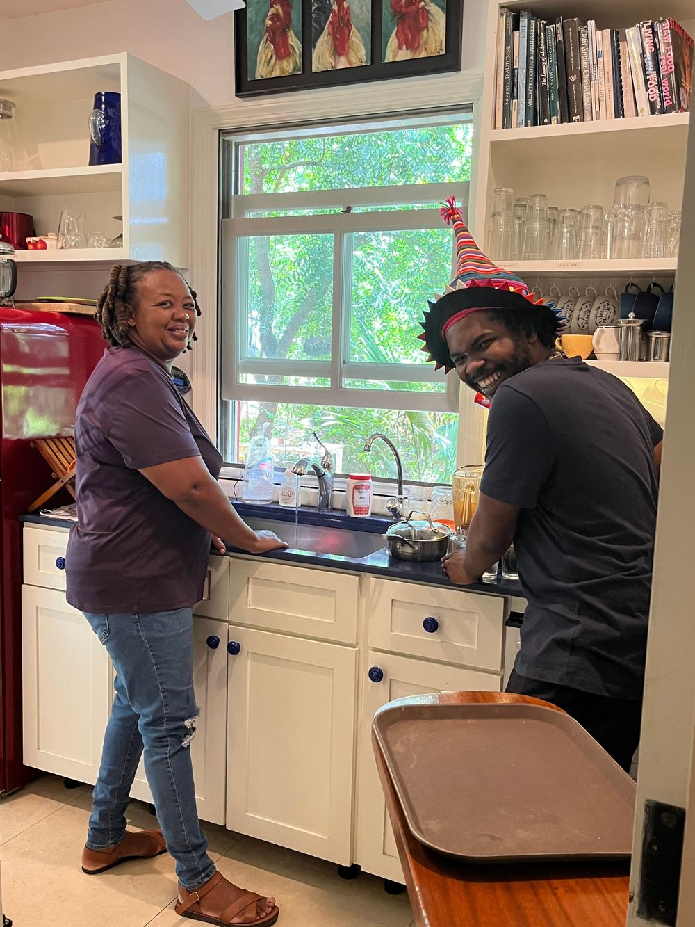 Two people smiling in a kitchen, one wearing a festive party hat, standing by the sink surrounded by shelves.