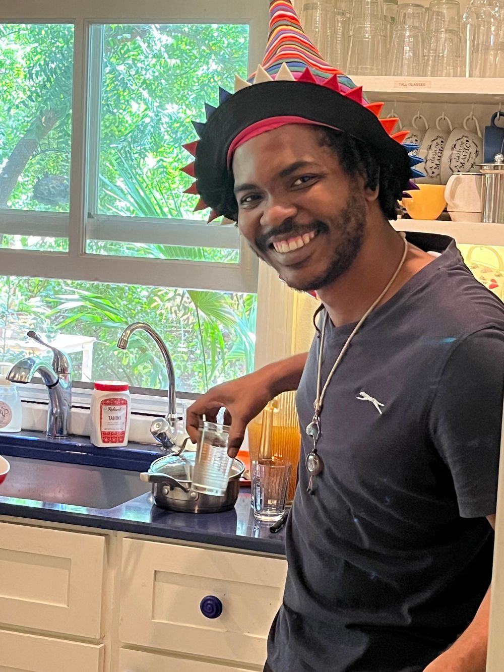 Smiling man wearing colorful party hat stands at kitchen sink, lush green trees visible through window behind him.