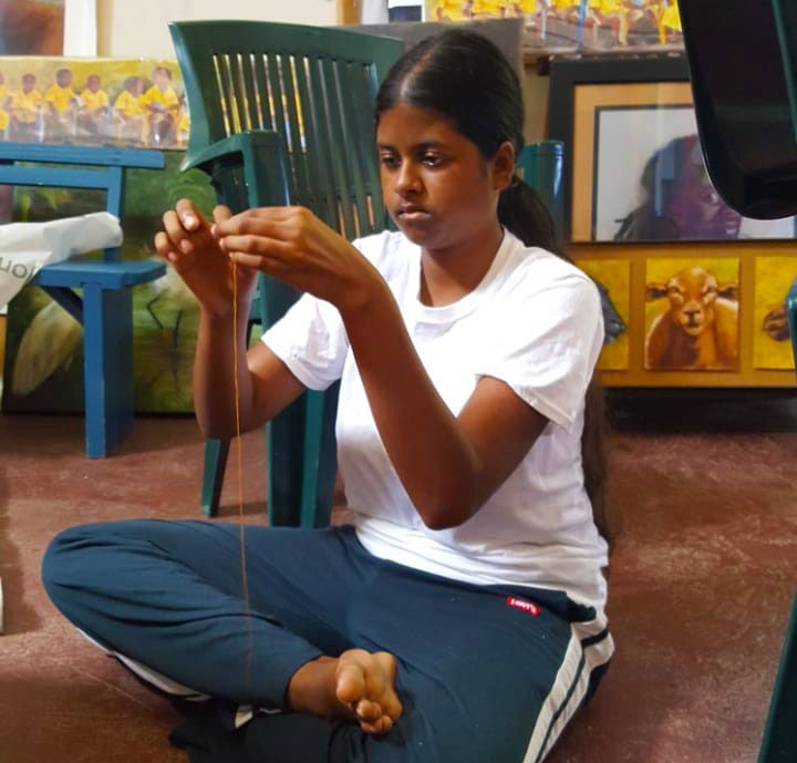 Young girl sitting cross-legged on floor, carefully threading a string with focused concentration indoors.