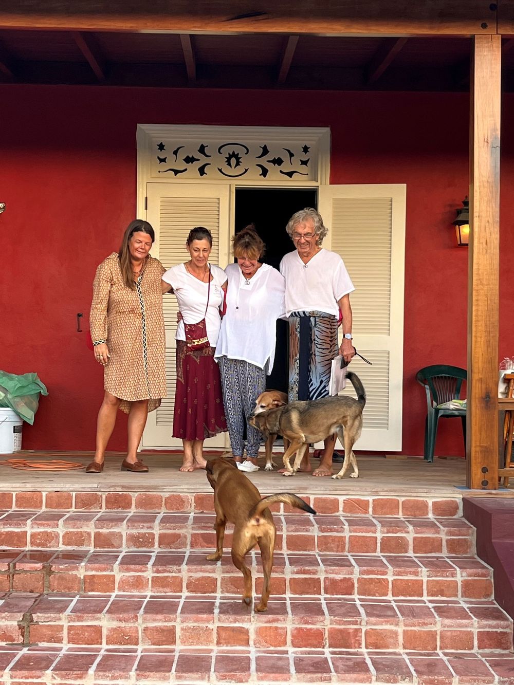 Four people pose on a red brick porch of a red building with two dogs in a sunny outdoor setting.
