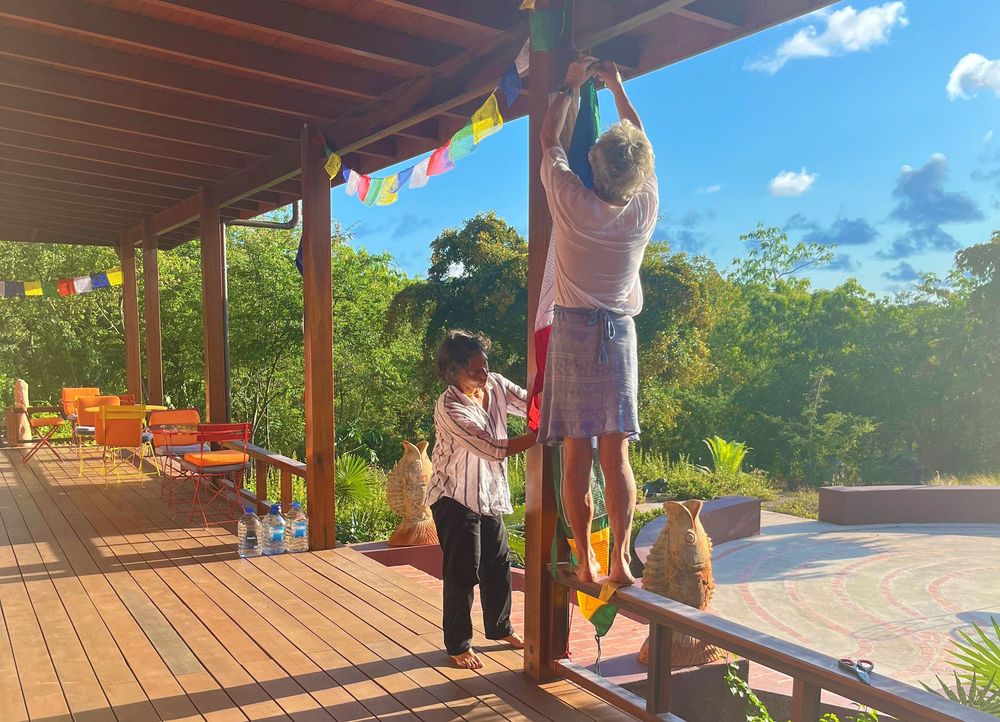 Two people hang colorful prayer flags on a wooden deck surrounded by lush tropical greenery under a bright blue sky.