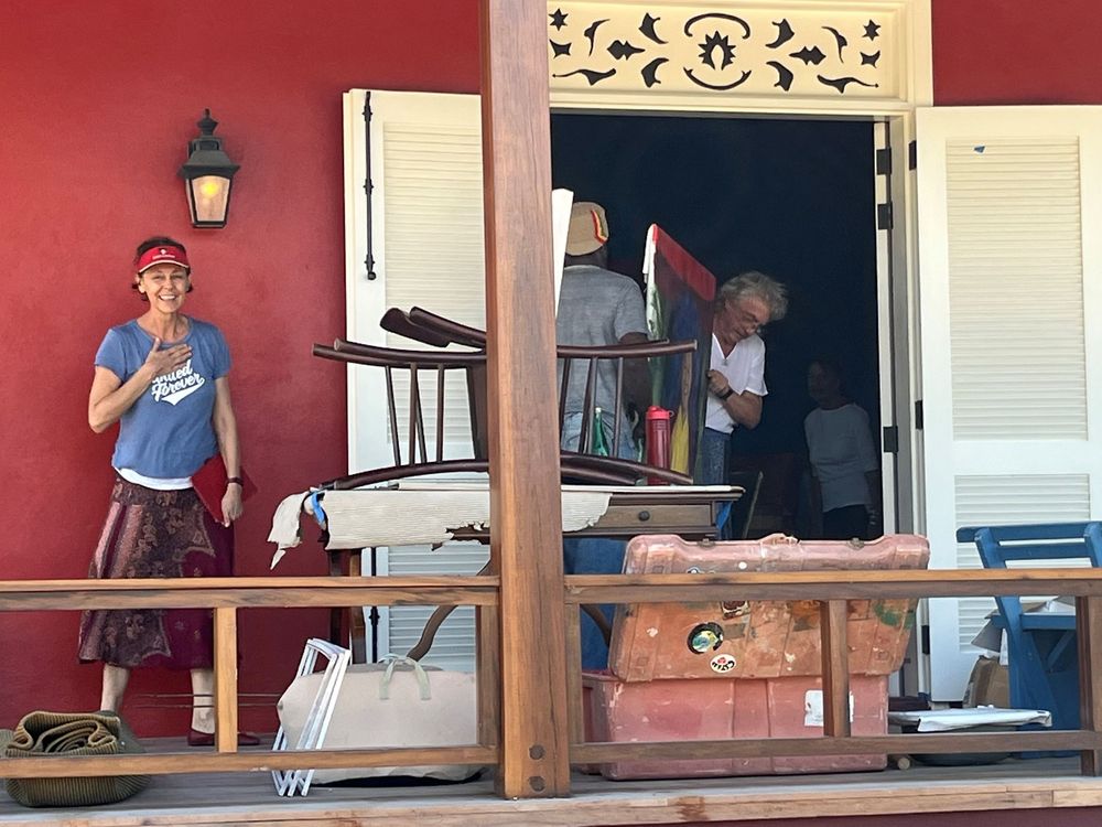 People moving furniture and belongings on a red building porch, with a woman standing outside observing the activity.
