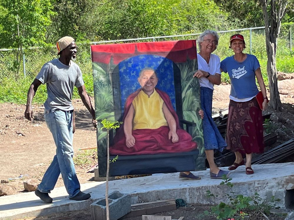 Three people outdoors holding a large portrait painting of a bald man in yellow robes against a blue background.