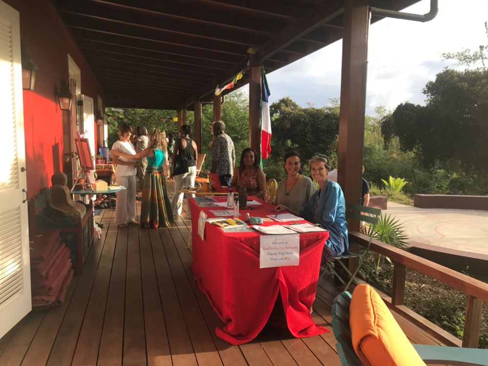 People gathered on a wooden deck with a red-draped table, flags, and lush greenery at an outdoor event.