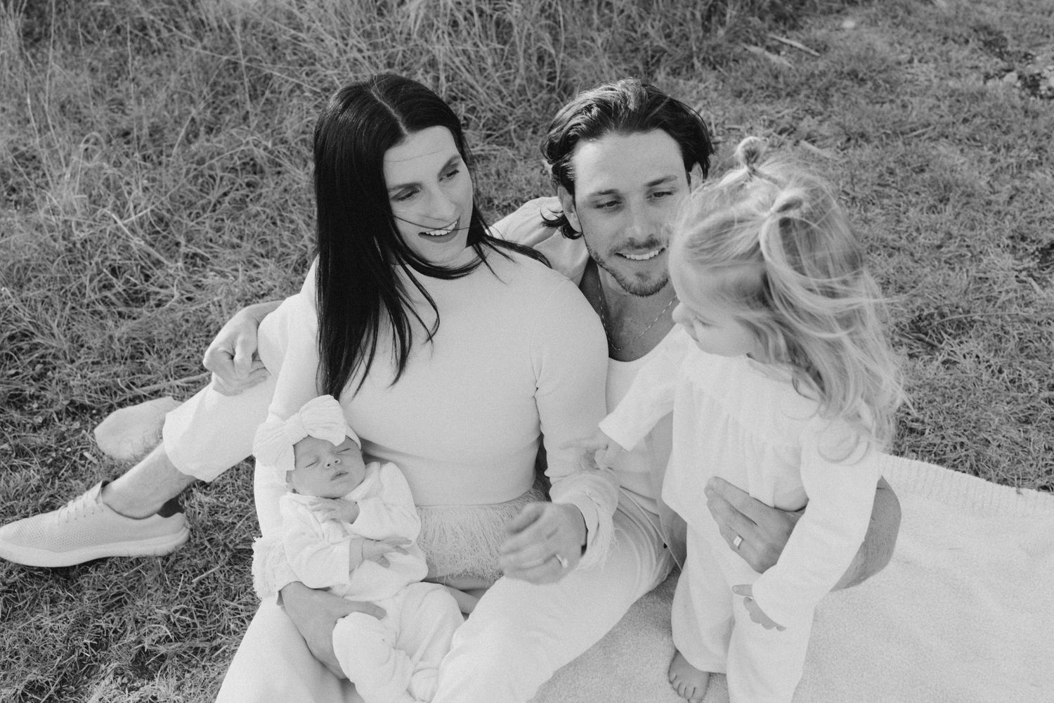 Black and white family photo of parents with baby and young child sitting on blanket in grass field.