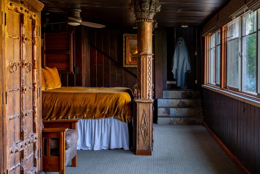Rustic bedroom with ornate wooden bed, golden satin bedding, carved pillar, and large windows in a dark wood-paneled room.