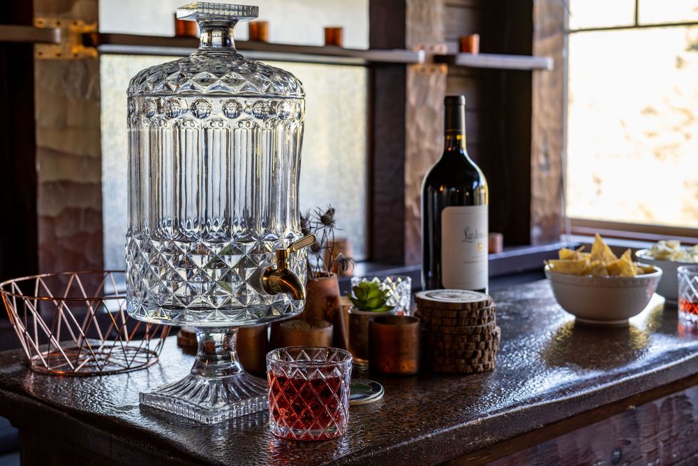 Elegant bar setup with crystal decanter, red wine bottle, cocktail glass, and snack bowls on a dark counter.