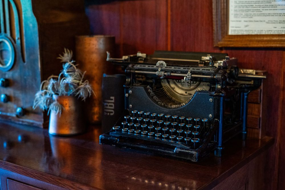 Vintage black typewriter on a mahogany desk beside a copper vase with dried plants in a classic study.