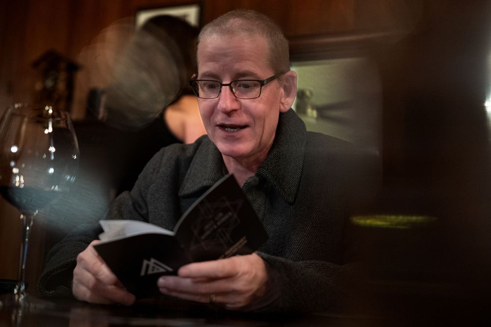 Man with glasses reading a book at a dimly lit table with a wine glass nearby.