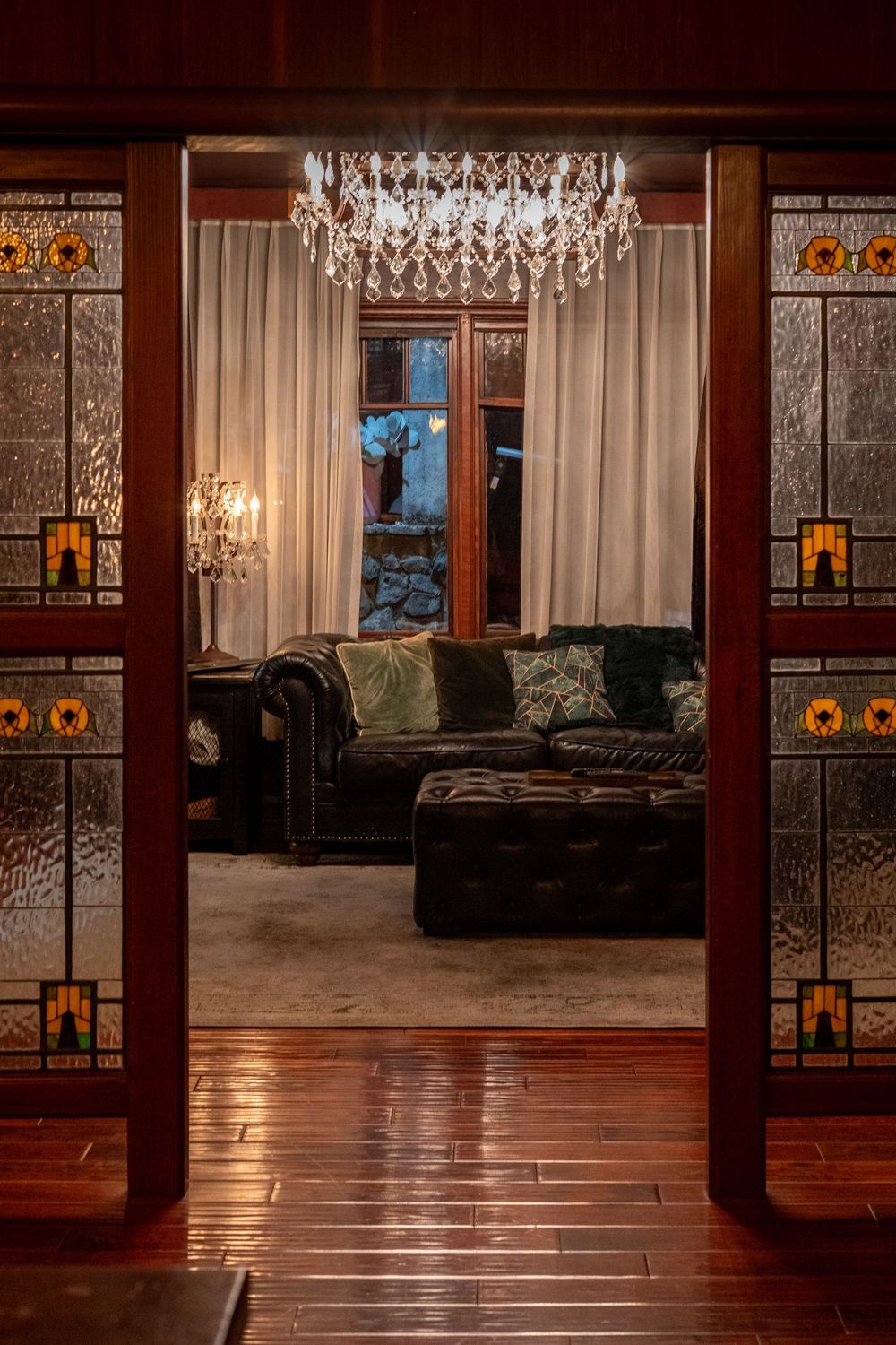 Elegant living room with crystal chandelier, Chesterfield sofa, stained glass doors, and hardwood floors in warm lighting.