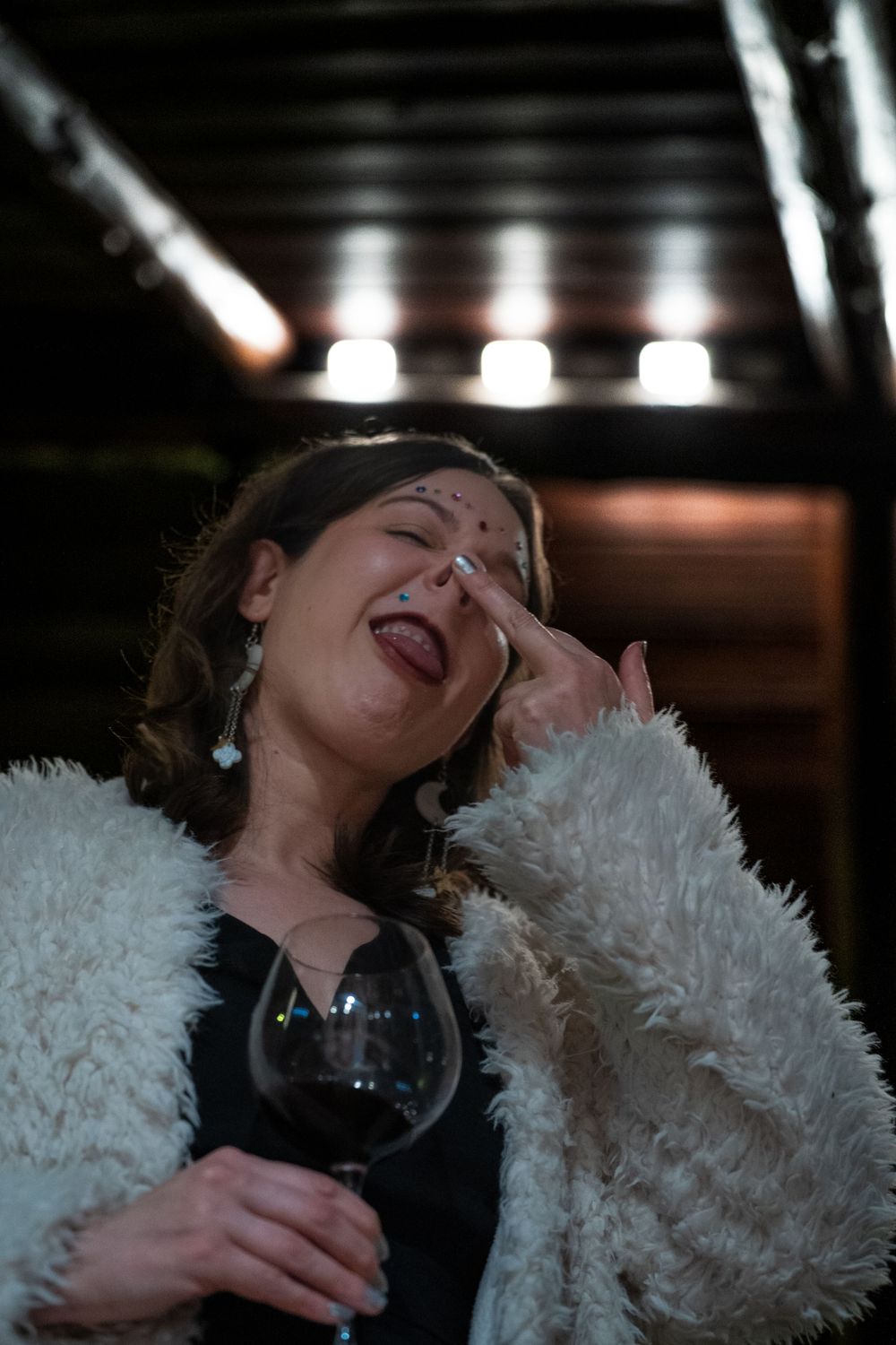 Woman in fluffy white coat laughing with eyes closed, holding a glass of red wine at a dimly lit evening event.