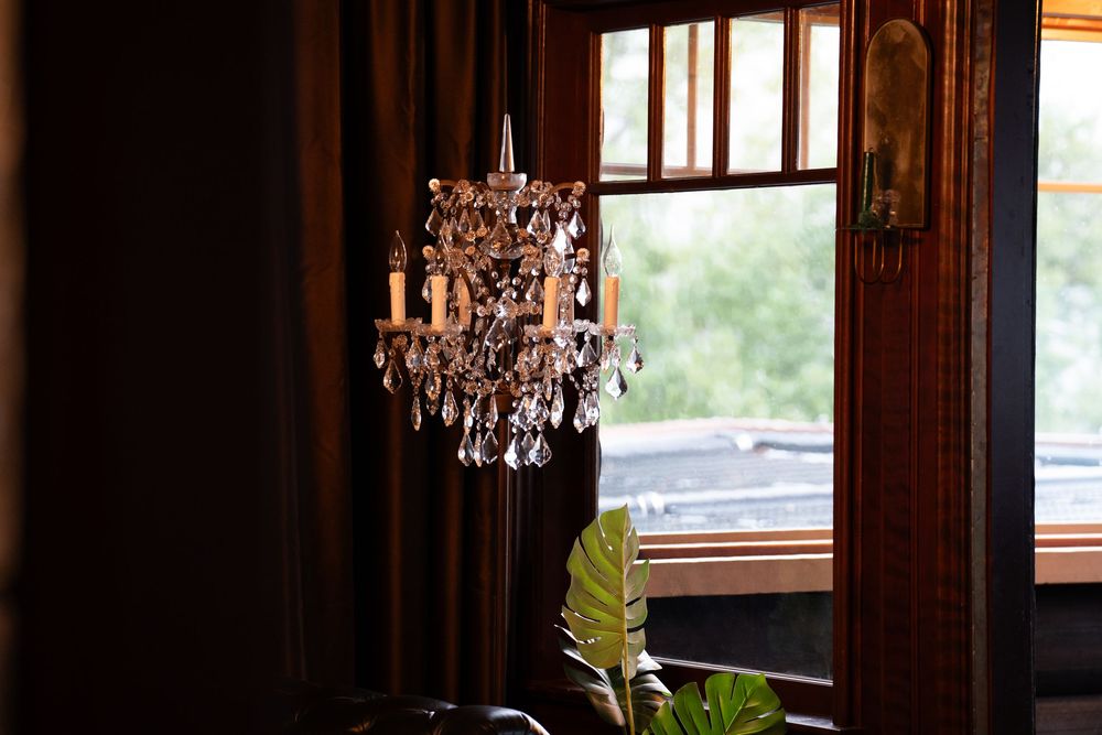 Elegant crystal chandelier hanging near a dark wood-framed window with green foliage visible outside in a vintage room.
