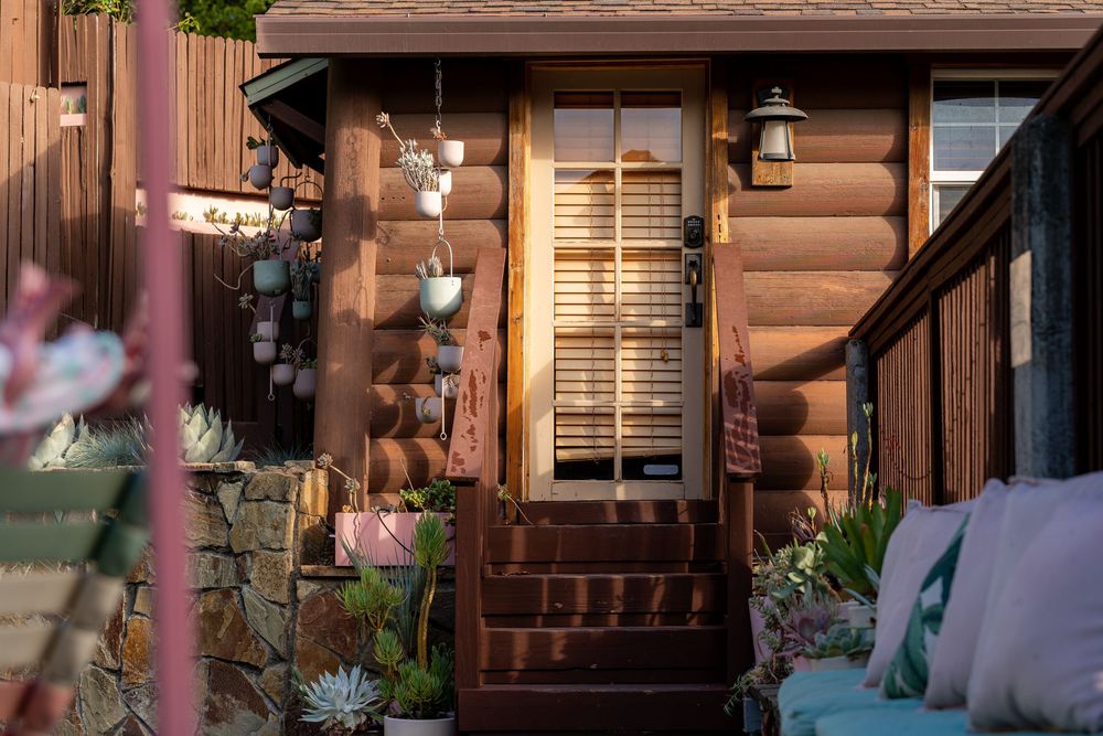Cozy log cabin entrance with warm glowing door, lantern, succulents, and wind chimes at golden hour.