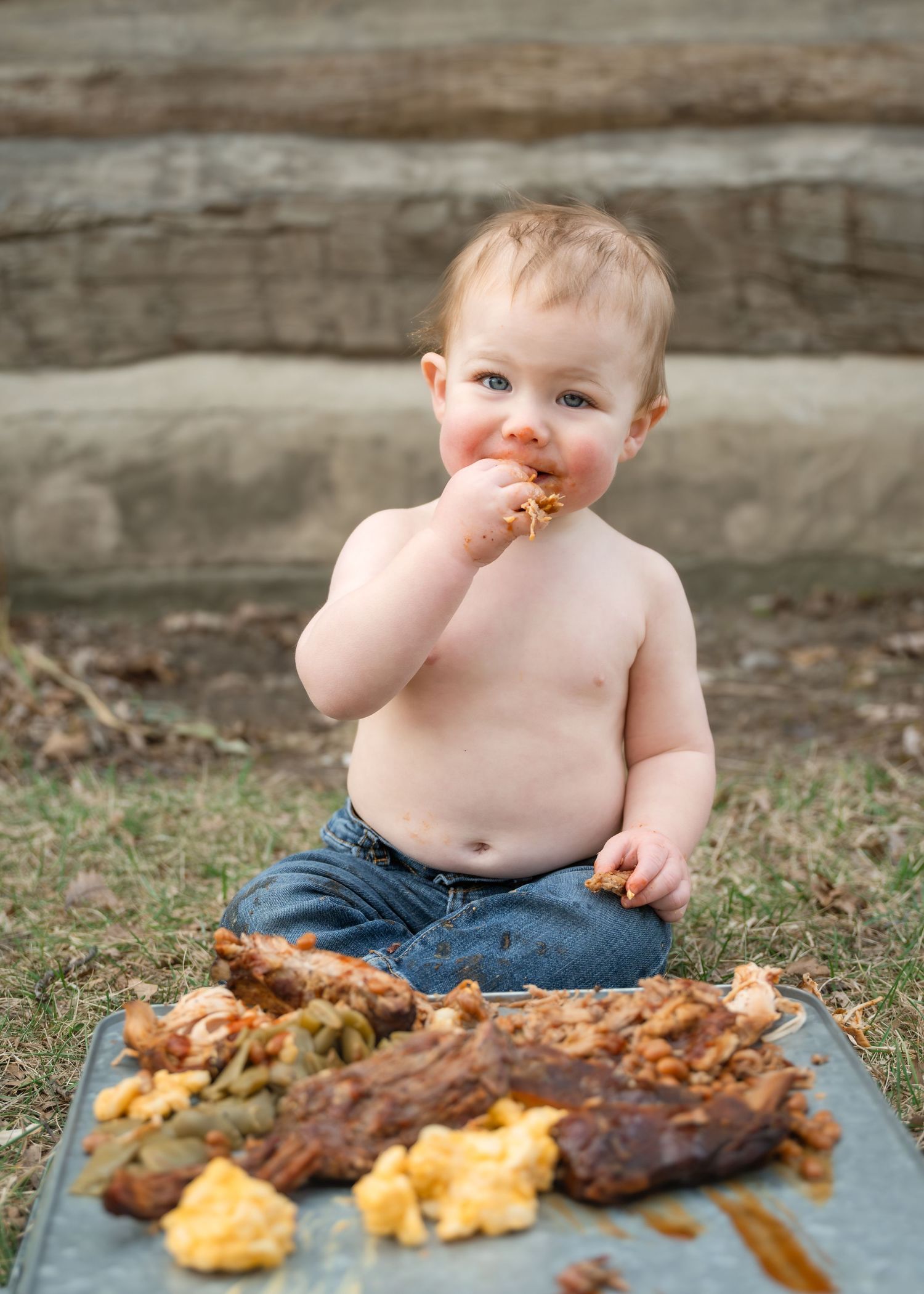 Shirtless baby boy sitting outdoors on a tray eating messy food with his hands, wearing jeans.
