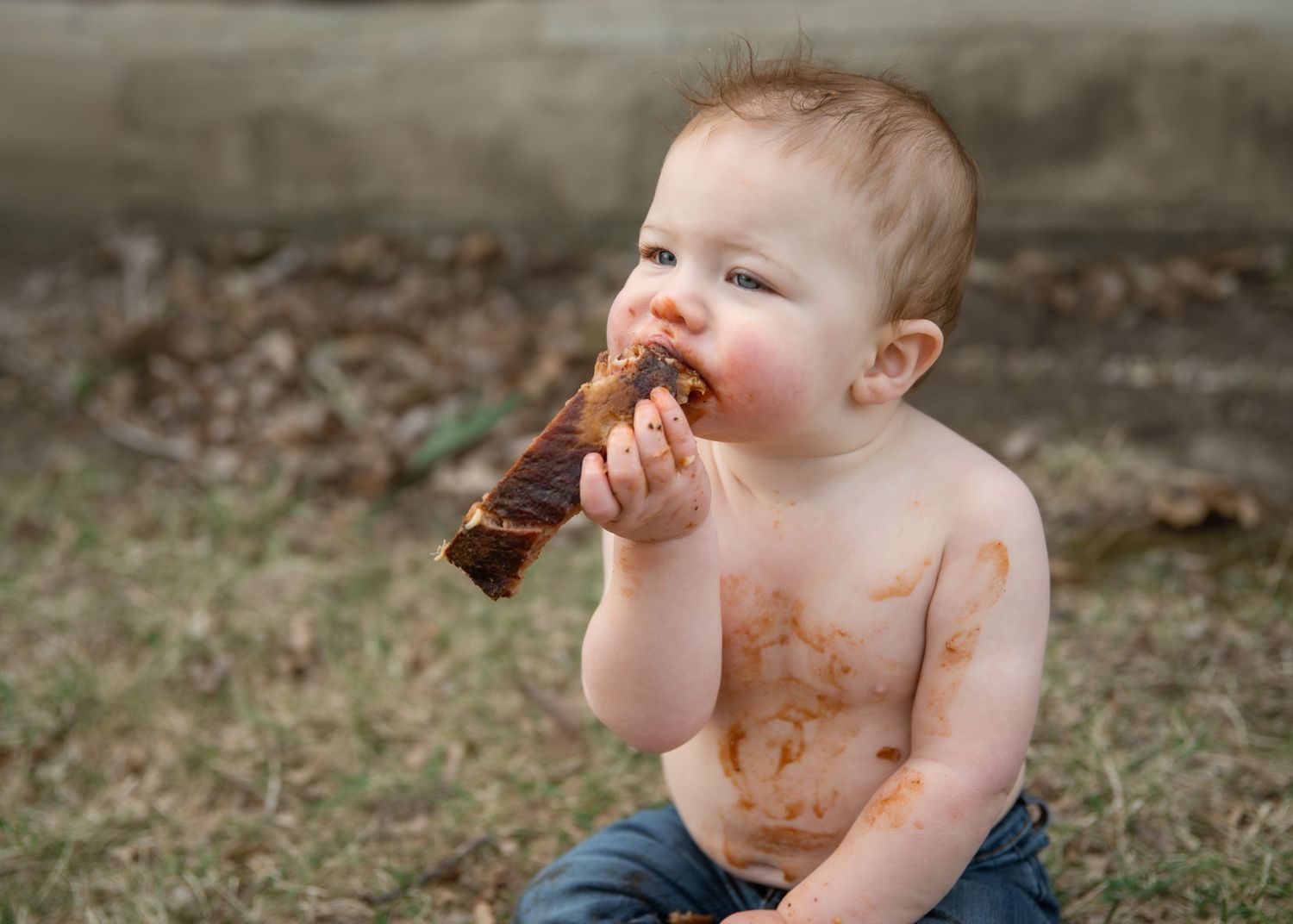 Messy baby eating BBQ ribs outdoors, covered in sauce, sitting on grass shirtless in jeans.